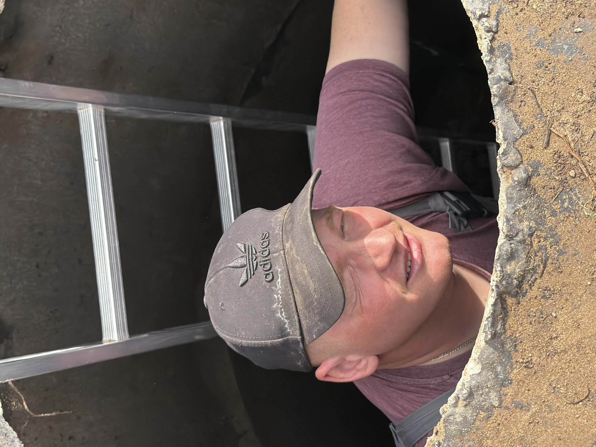 A man wearing a maroon t-shirt and a dusty gray Adidas baseball cap is partially emerged from a round, concrete hole in the ground. A silver metal ladder is visible inside the hole. The setting is outside. This image does not provide enough detail to determine the location in the city of Chaikovsky, Russia.