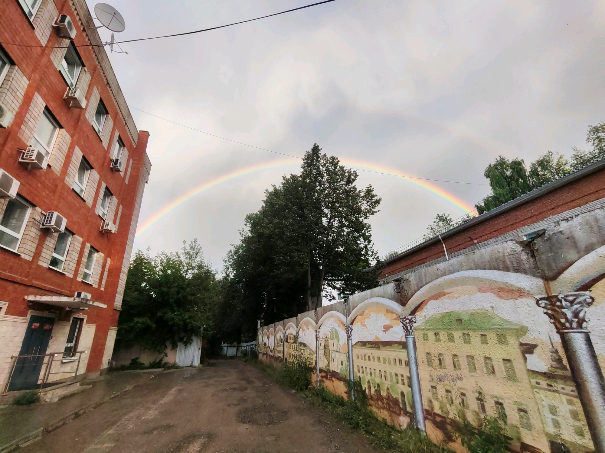 The image shows a view of a street in Izhevsk, Russia, with a prominent rainbow stretching across a cloudy sky. On the left side of the frame, a red brick apartment building with white-framed windows and multiple air conditioning units is visible. A satellite dish is mounted on the roof of this building. A dirt path leads away from the building, bordered on the right by a long, arched mural depicting historical buildings. Trees line the top of the wall behind the mural and further into the background. The sky is overcast with a visible rainbow.