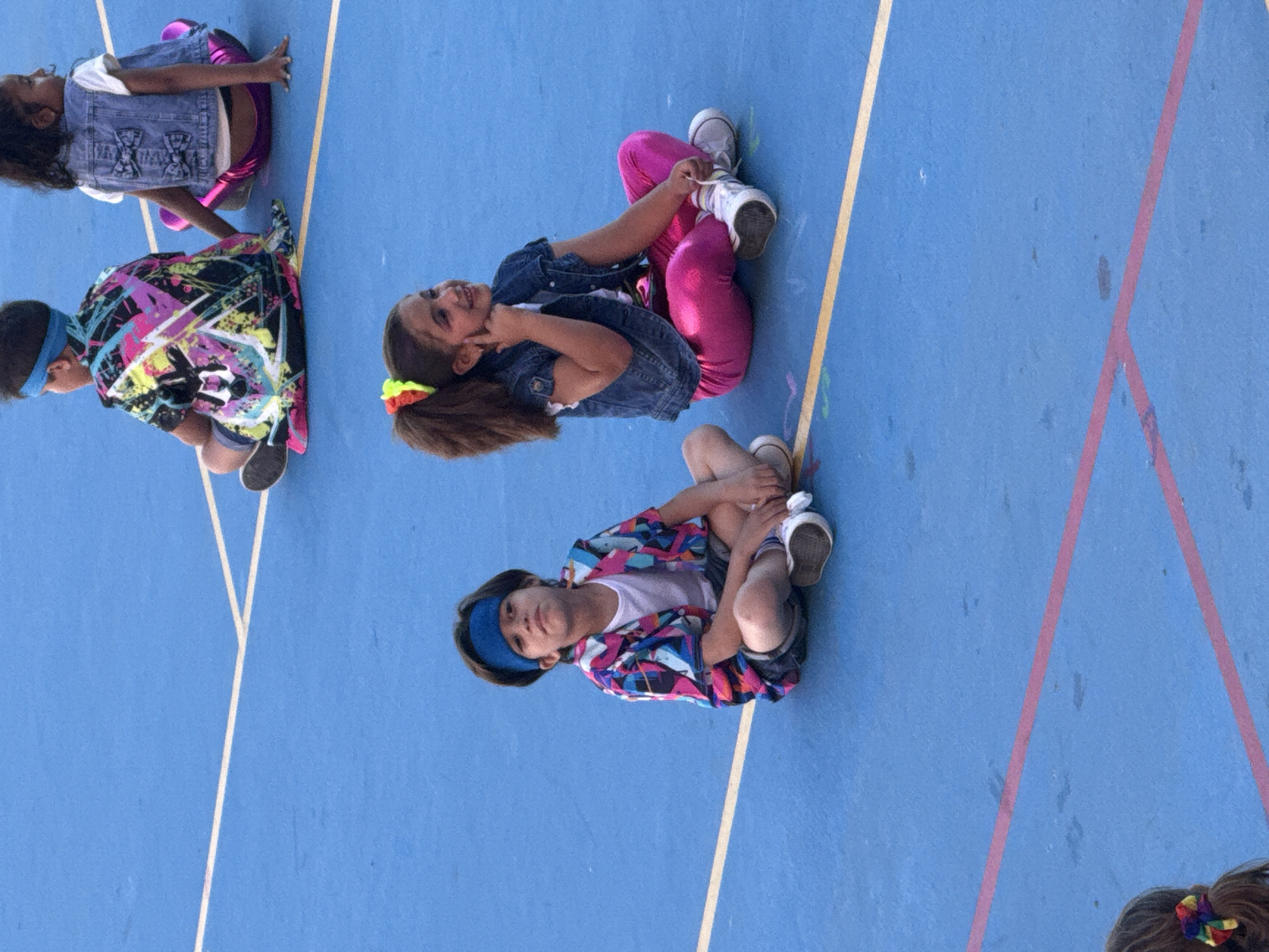 The image depicts four children sitting on a blue painted surface marked with yellow and pink lines. They appear to be arranged in a somewhat staggered formation, with one child each towards the top, center, and bottom of the frame, and one more to the left.
The child at the top of the frame is wearing a denim vest over a white shirt and metallic purple leggings. The child to the left is wearing a brightly colored shirt with a geometric pattern. The child in the center is wearing a denim jacket and bright pink leggings. The child at the bottom is wearing a colorful jacket over a white shirt. All the children are wearing shoes. They are posed in what looks like different seated positions.
The location appears to be outdoors on what might be a painted play surface, possibly in an urban area of an unknown city. The composition of the photograph suggests the picture was taken from an elevated perspective.