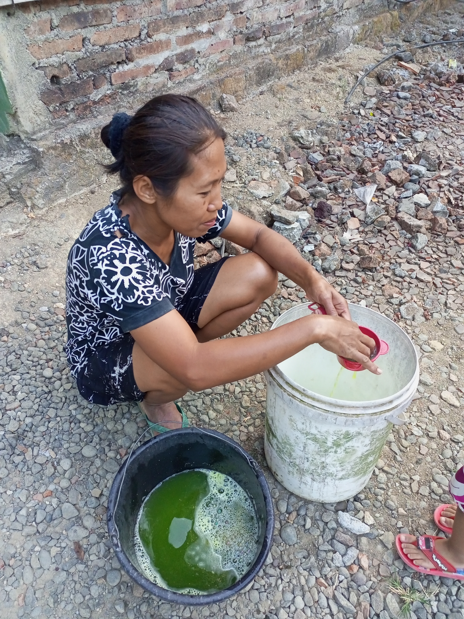 A woman of Southeast Asian descent, with dark hair pulled back in a bun and wearing a black and white patterned short-sleeved shirt and black shorts, crouches on a gravelly ground near a brick wall. She is using a red sieve to pour liquid from a red cup into a white bucket containing a light-colored liquid. Next to the white bucket is a black bucket filled with green, soapy water. Her feet are bare, clad in green flip-flops. The environment appears to be outdoors, with a rough, rocky ground surface and a weathered brick wall in the background. The scene is located in Nanggulan, Indonesia.