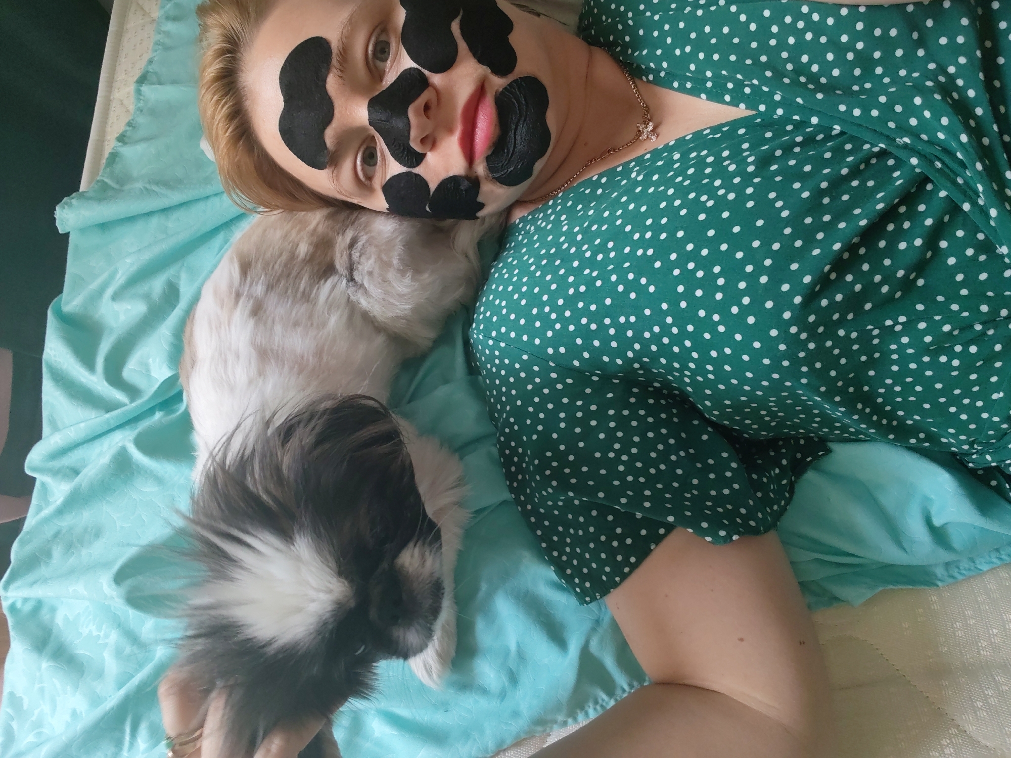 In Izhevsk, Russia, a fair-skinned woman with blonde hair lies on her back on a pale blue textured blanket, with a small, fluffy dog nestled beside her. The woman is wearing a green polka-dot dress and has black charcoal mask patches applied to her face. The dog has light brown and white fur. The scene is captured from a top-down perspective.