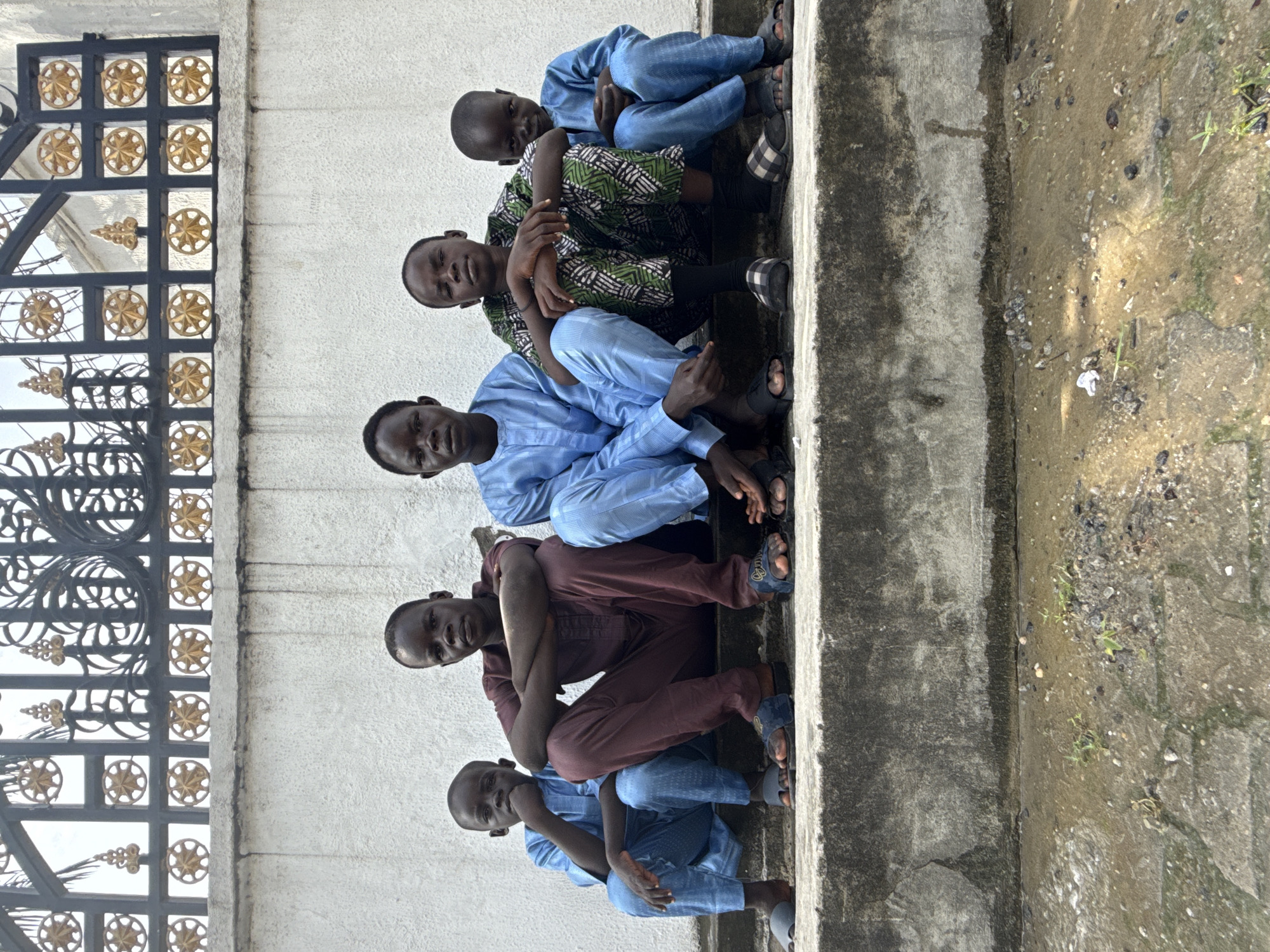 Five males are seated on concrete steps next to a wall and an ornate metal gate, likely in Ado, Nigeria. The males are wearing various colored clothing and sandals. The environment shows signs of moisture and vegetation growth near the concrete steps.