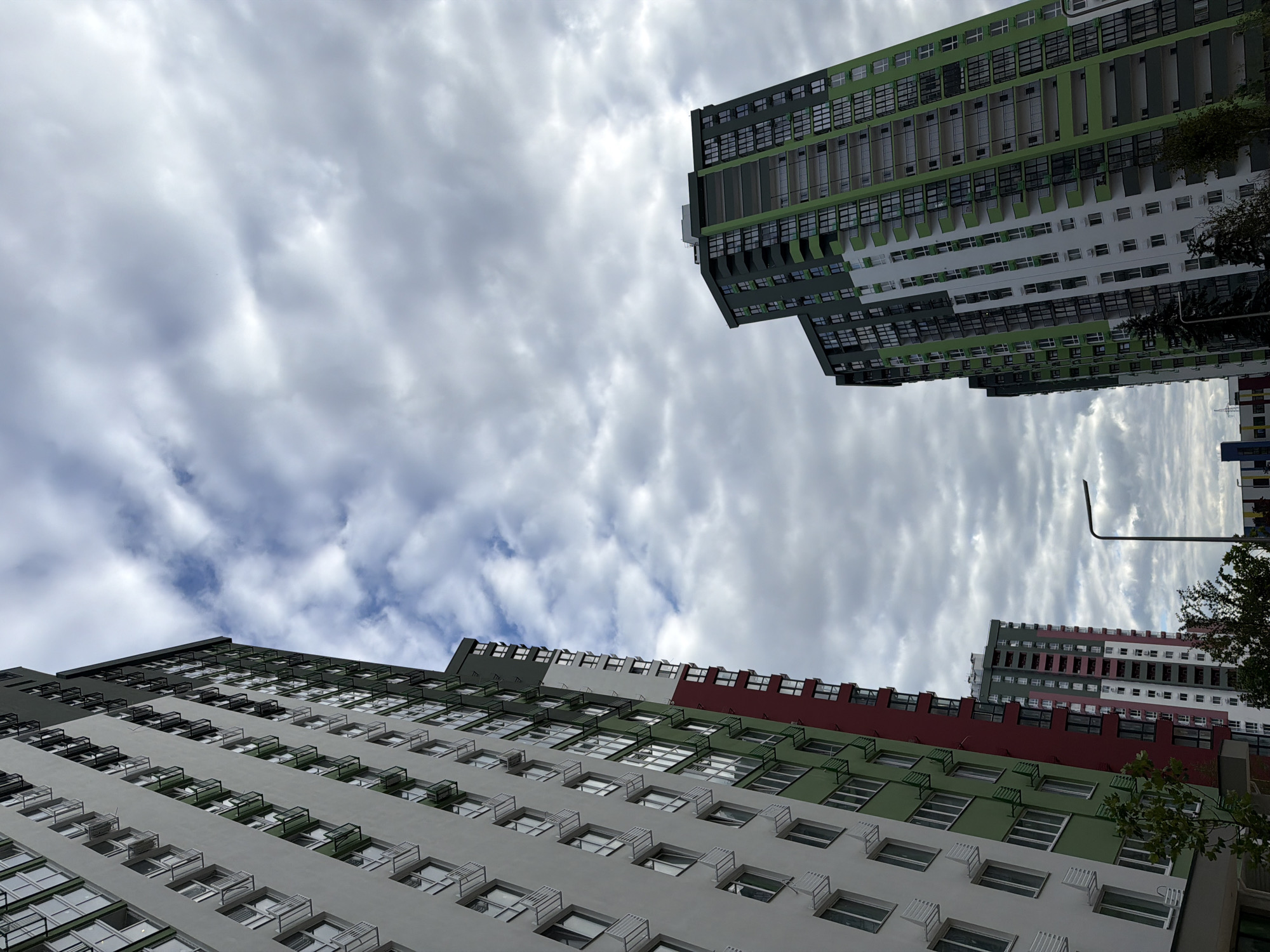 In Kyiv, Ukraine, a low-angle shot captures a cluster of modern, multi-story residential buildings against a cloudy sky. The buildings are characterized by their distinct color blocking, featuring sections of white, green, and red facades. Numerous windows, some with visible balconies or air conditioning units, punctuate the exteriors. One building on the left is predominantly white with vertical green and red accents. To its right, a taller building displays a striking pattern of green, white, and red. Further to the right, another tall structure showcases a design dominated by green and white sections, with balconies visible on its upper floors. A street lamp stands in the lower right portion of the frame, and trees are visible at the base of the buildings. The sky is filled with textured, grey clouds, suggesting overcast weather. The scene is static, with no people or discernible activity.