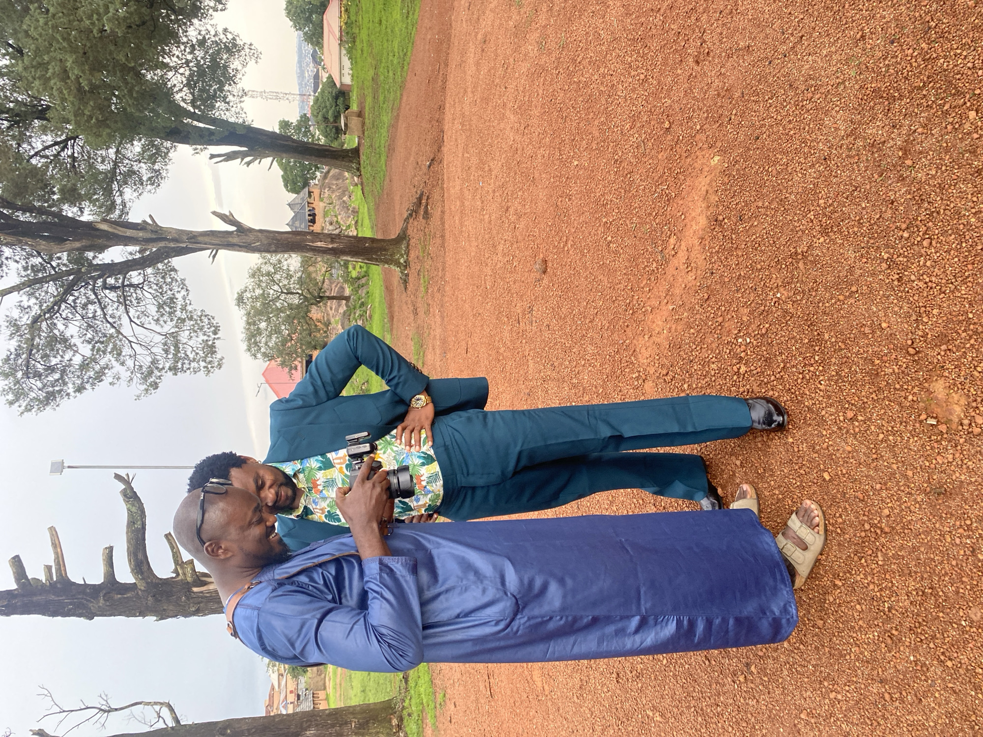 Two men are interacting outdoors, with one man holding a camera and appearing to take a picture or record something. The man holding the camera is wearing a blue robe and sandals. The other man is wearing a teal suit with a floral shirt underneath and dress shoes. He is looking down and smiling. The scene is set in a park-like area with a dirt path, grass, and trees. In the background, there are some buildings and structures, suggesting a semi-urban or rural environment. The sky is overcast, indicating a cloudy day. The man in the suit is wearing a watch. The overall mood seems relaxed and friendly. The location context is Jos, Nigeria.