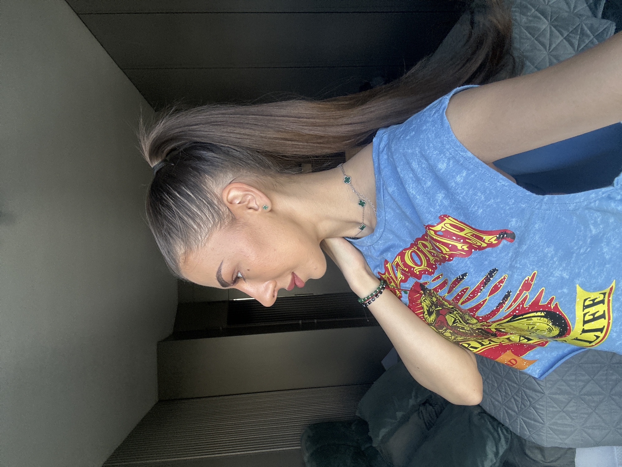 A young woman with long, dark brown hair pulled into a high ponytail is photographed from the side in a room. She is wearing a light blue t-shirt with a graphic print. She has on a delicate necklace with green clover-shaped charms and a bracelet on her left wrist. Her right arm is bent at the elbow, with her hand resting on her neck. The background shows a bed with dark green pillows and a gray quilted bedspread, and a closet with dark wooden doors. The lighting suggests it is daytime, with light coming from a window that is not visible in the frame. The room appears to be a bedroom, possibly in an apartment in Sofia, Bulgaria.