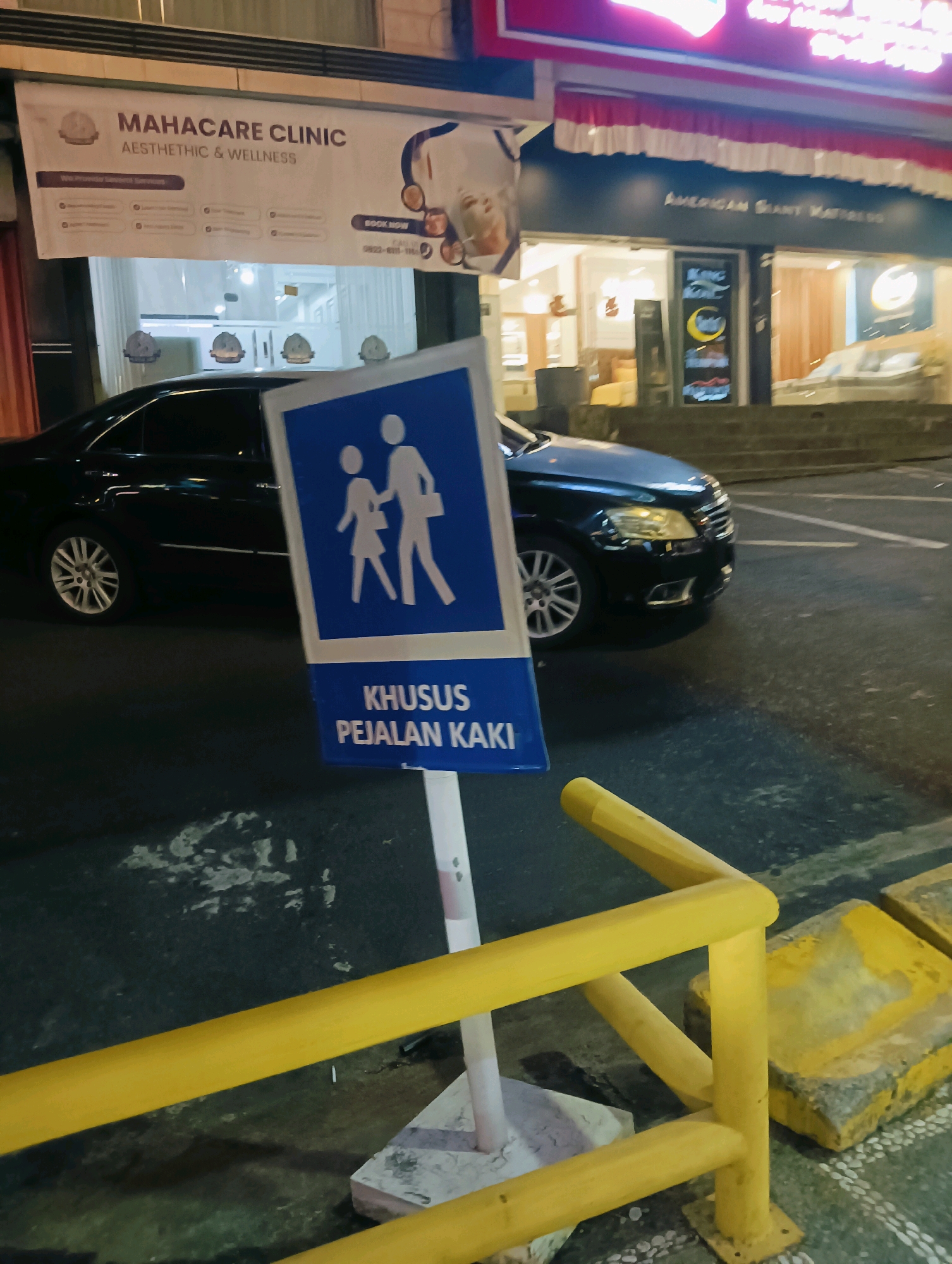 In Batam, Indonesia, at night, a pedestrian crossing sign is prominently displayed. The sign, a blue rectangle with white figures of two people walking, signifies a pedestrian area. Below this graphic, text in Indonesian reads "KHUSUS PEJALAN KAKI," translating to "FOR PEDESTRIANS." The sign stands on a white pole, anchored in the asphalt. A yellow metal barrier frames the lower right corner of the image, partially obscuring concrete blocks and the textured road surface. In the background, a dark-colored sedan is parked, its headlights illuminating the wet asphalt. Behind the car, storefronts are visible, including "MAHACARE CLINIC AESTHETIC & WELLNESS" on a banner, with the clinic's interior illuminated. Further down the street, another storefront displays "AMERICAN GIANT MATTRESS." The overall scene suggests an urban commercial area at dusk or night.