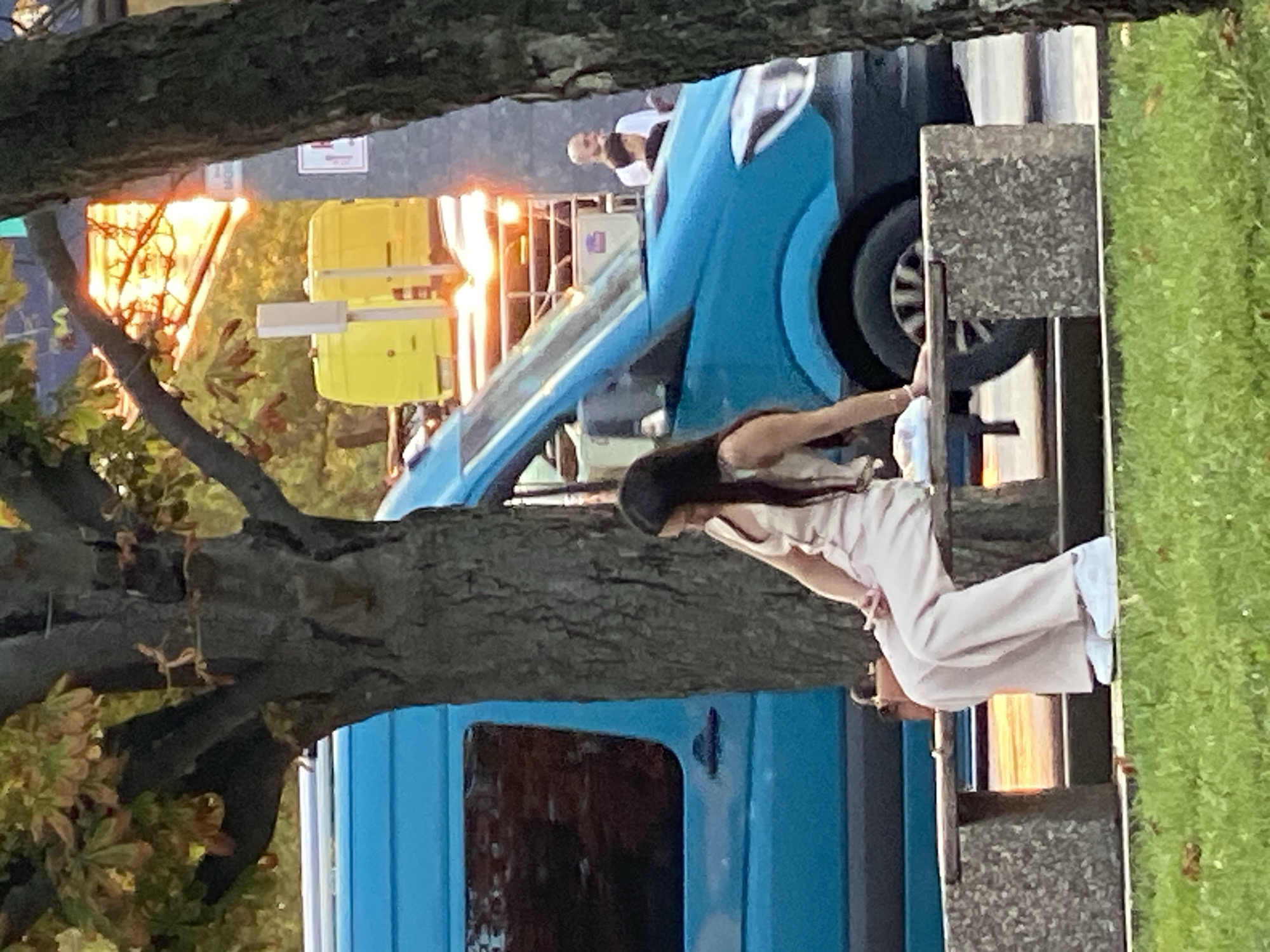 A young woman with long dark hair is sitting on a concrete and wooden bench, her legs are extended and her arms are resting on her legs. She is wearing a light-colored matching set of a camisole and wide-leg pants, and white sneakers. A brown handbag rests on the bench next to her. In the background, a bright blue van is parked. Beyond the van, two yellow boxes are visible, along with the warm glow of setting sun. A person wearing a white shirt is standing near the van. The scene appears to be in a public area of Chișinău, Moldova, possibly a park or a street with benches and a park nearby.