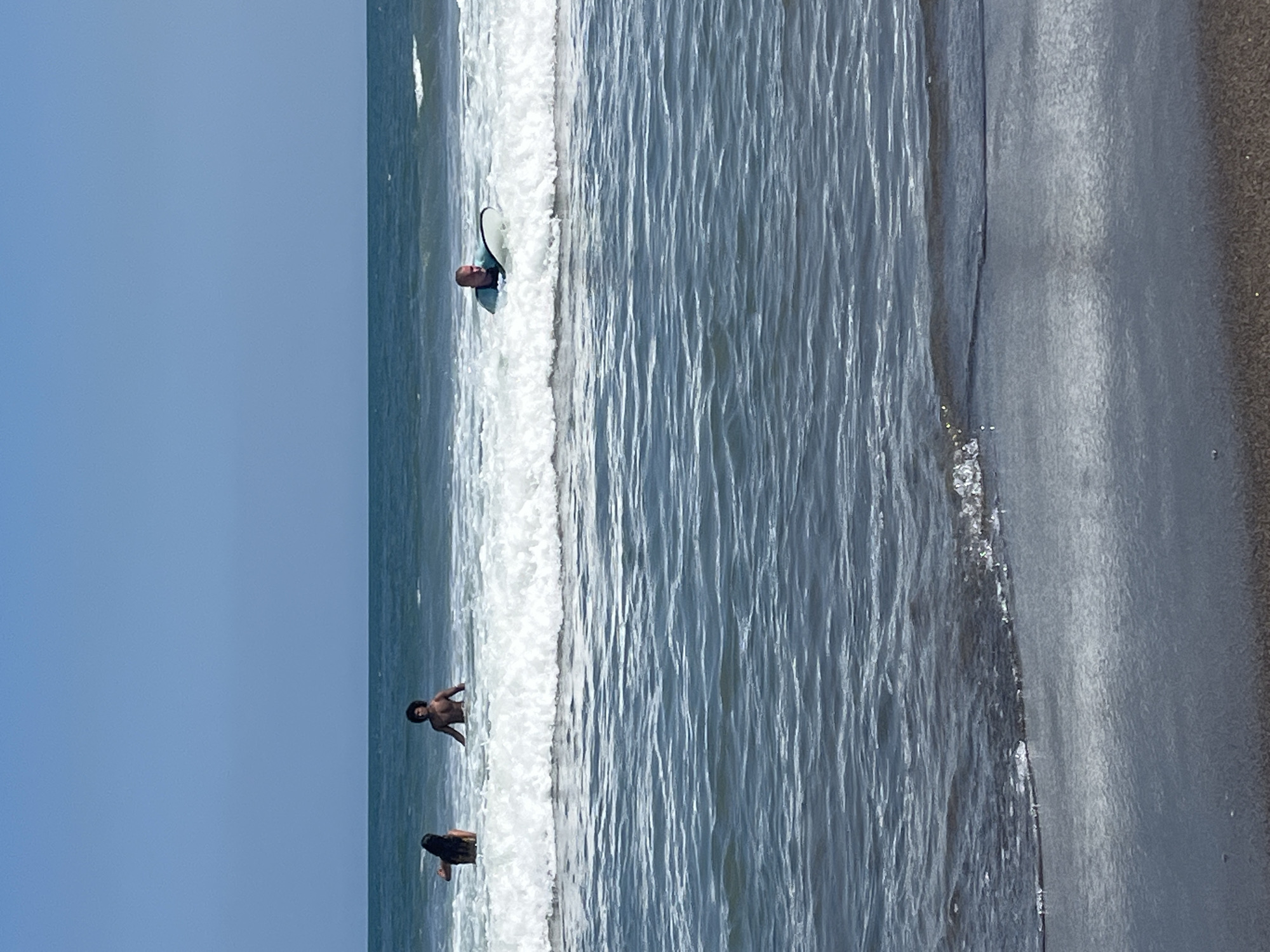 In North Kuta, Indonesia, on a clear day with a blue sky, three people are in the ocean. In the distance, two individuals are standing in the water, with the person on the left appearing to be a woman with dark hair. To the right of them, a man with curly dark hair is also standing in the water. Closer to the foreground, a person is lying on a surfboard in the ocean, partially submerged in a wave. The water is a deep blue, and the waves are breaking with white foam. The beach in the foreground is dark and sandy.