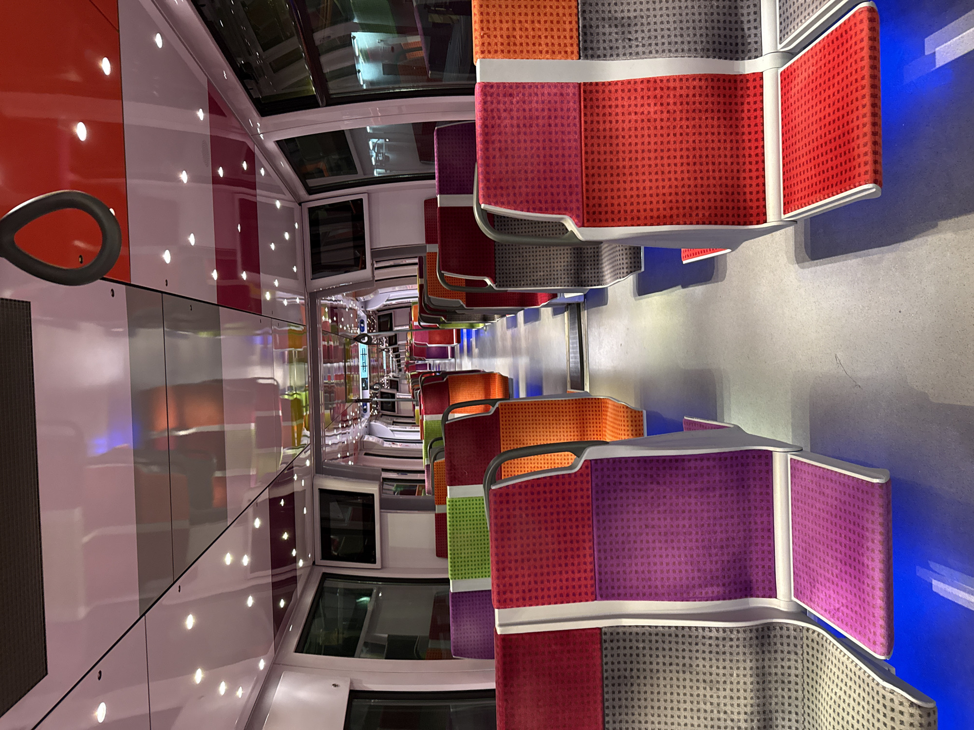 The image shows the interior of a train, likely a suburban train, in Saint-Brice-sous-Forêt, France. The train car features rows of seats upholstered in a variety of bright colors, including red, orange, green, and purple, each paired with gray fabric on the lower section of the seatback. The seats are arranged facing each other along the length of the car.
The walls of the train car are white with vertical red and gray bands, and are punctuated by evenly spaced circular lights. There are also square windows, some of which have integrated screens. The floor is a light gray color. The angle of the photo creates a sense of perspective, with the rows of seats converging in the distance.