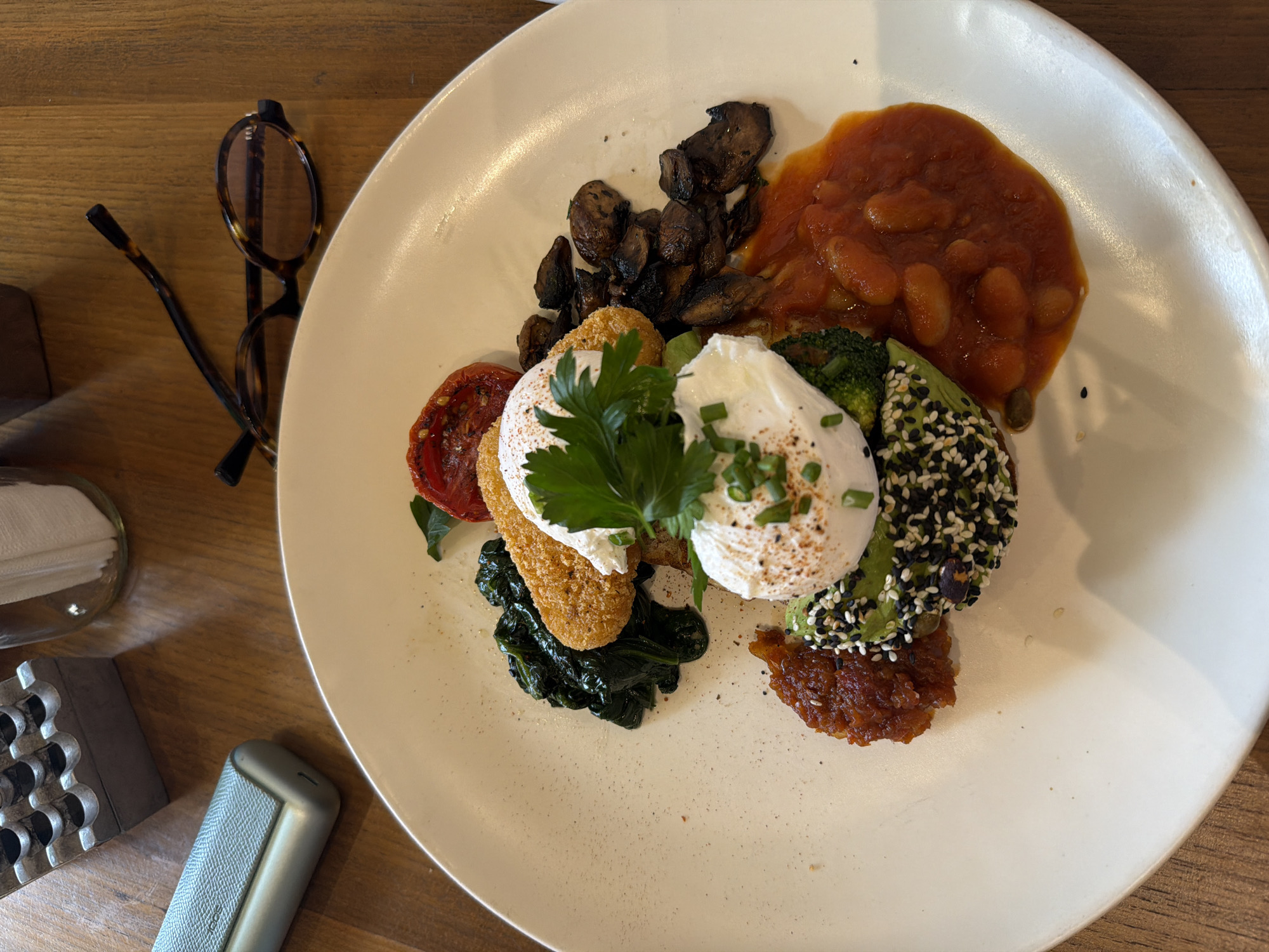 A close-up, overhead shot captures a white plate of food on a wooden table, with out-of-focus elements of eyeglasses and a silver electronic cigarette case in the background. The plate holds a breakfast dish featuring a poached egg atop avocado coated with black and white sesame seeds, surrounded by baked beans in tomato sauce, sautéed mushrooms, wilted spinach, a breaded cutlet, and a halved roasted tomato. The scene is set in a cafe in North Kuta, Indonesia.