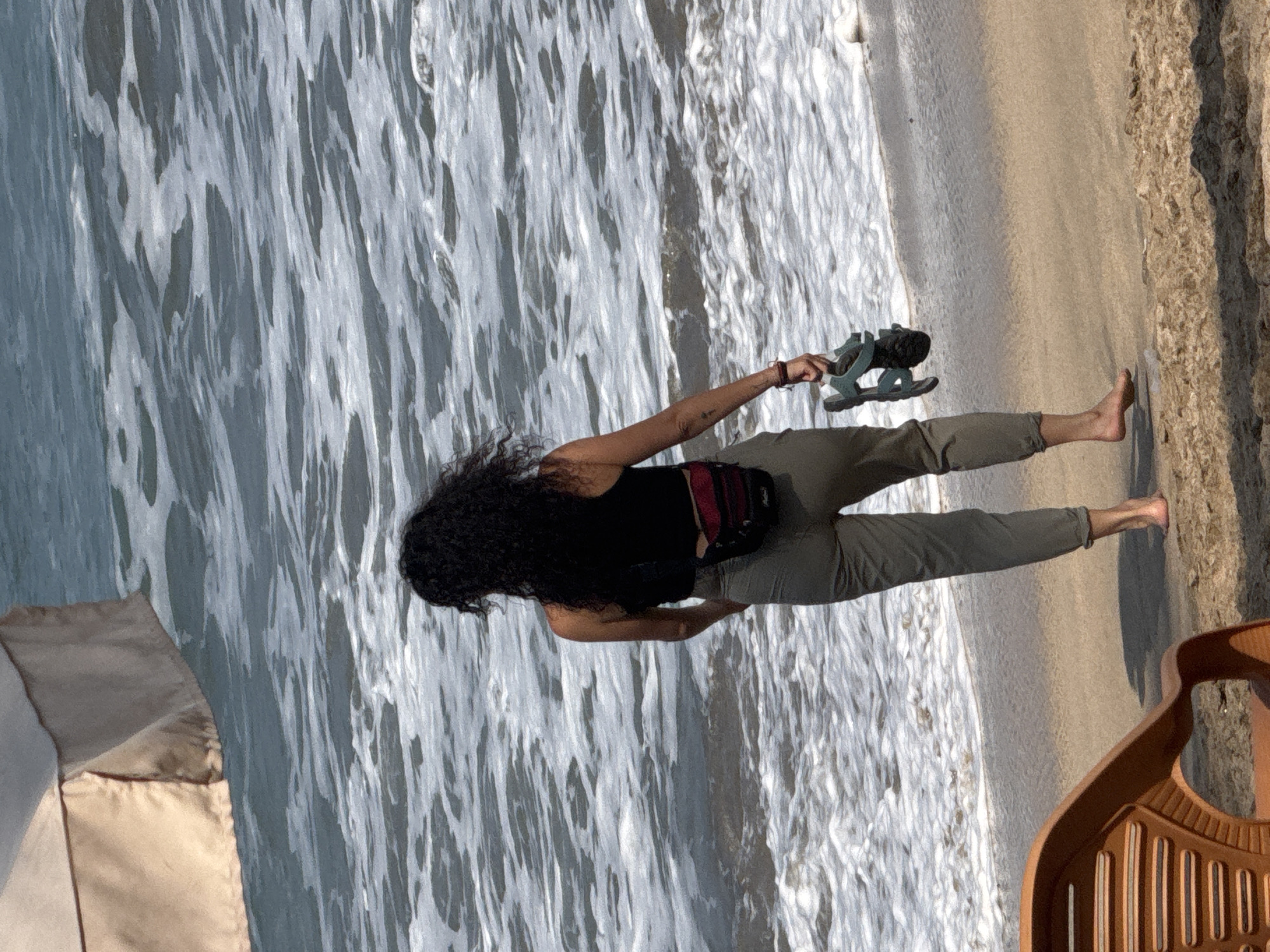A woman with curly dark hair stands on a sandy beach in Kuta, Indonesia, facing the ocean. She is wearing a black tank top and olive-green pants, with a small red and black fanny pack around her waist. In her right hand, she holds a pair of dark-colored sandals. The ocean behind her is blue with white waves. In the foreground, the edge of an orange plastic beach chair is visible. A beige umbrella is partially visible in the upper left corner of the image.