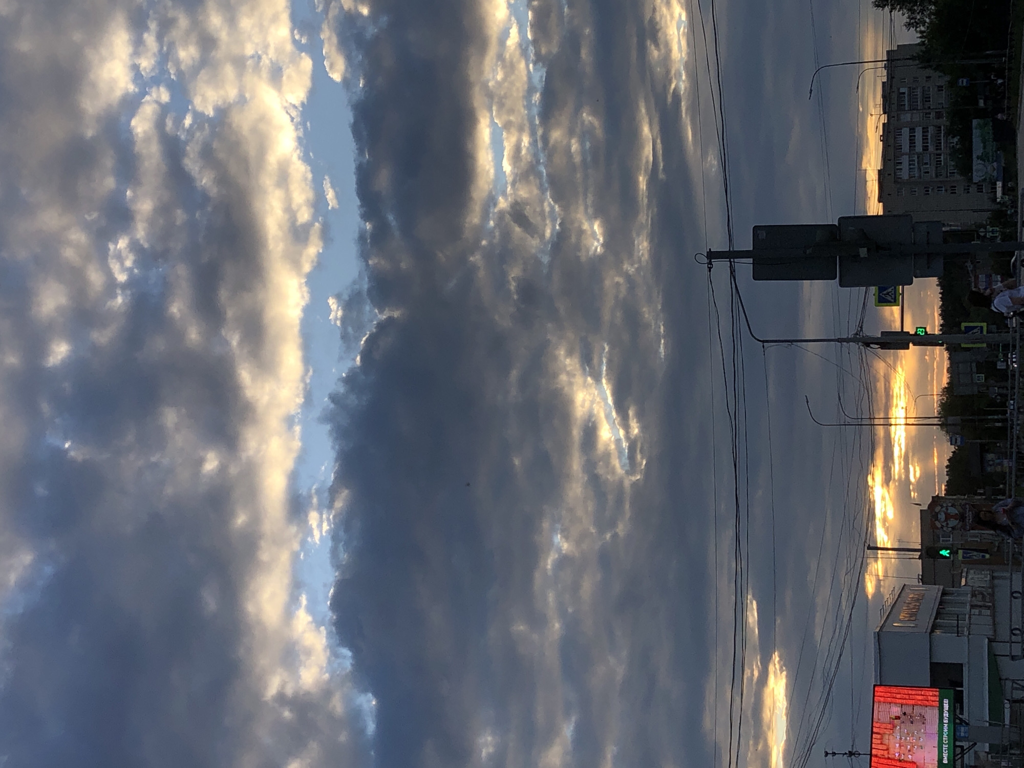 The image captures a cloudy sky over the city of Chaikovsky, Russia, likely during sunset or sunrise. The sky is filled with dramatic clouds, with shafts of golden light breaking through, illuminating patches of blue sky and casting an ethereal glow on the cloud formations. Below the sky, the urban landscape is visible, with buildings, streetlights, traffic signals, and electrical wires creating a layered effect. In the foreground, a pedestrian crossing is indicated by a green traffic light. Several people are present, walking on the sidewalk or near the road, their figures relatively small against the vastness of the sky and urban structures. One woman in the foreground is holding what appears to be a drink. The scene conveys a sense of everyday life unfolding under a dynamic and visually striking sky.