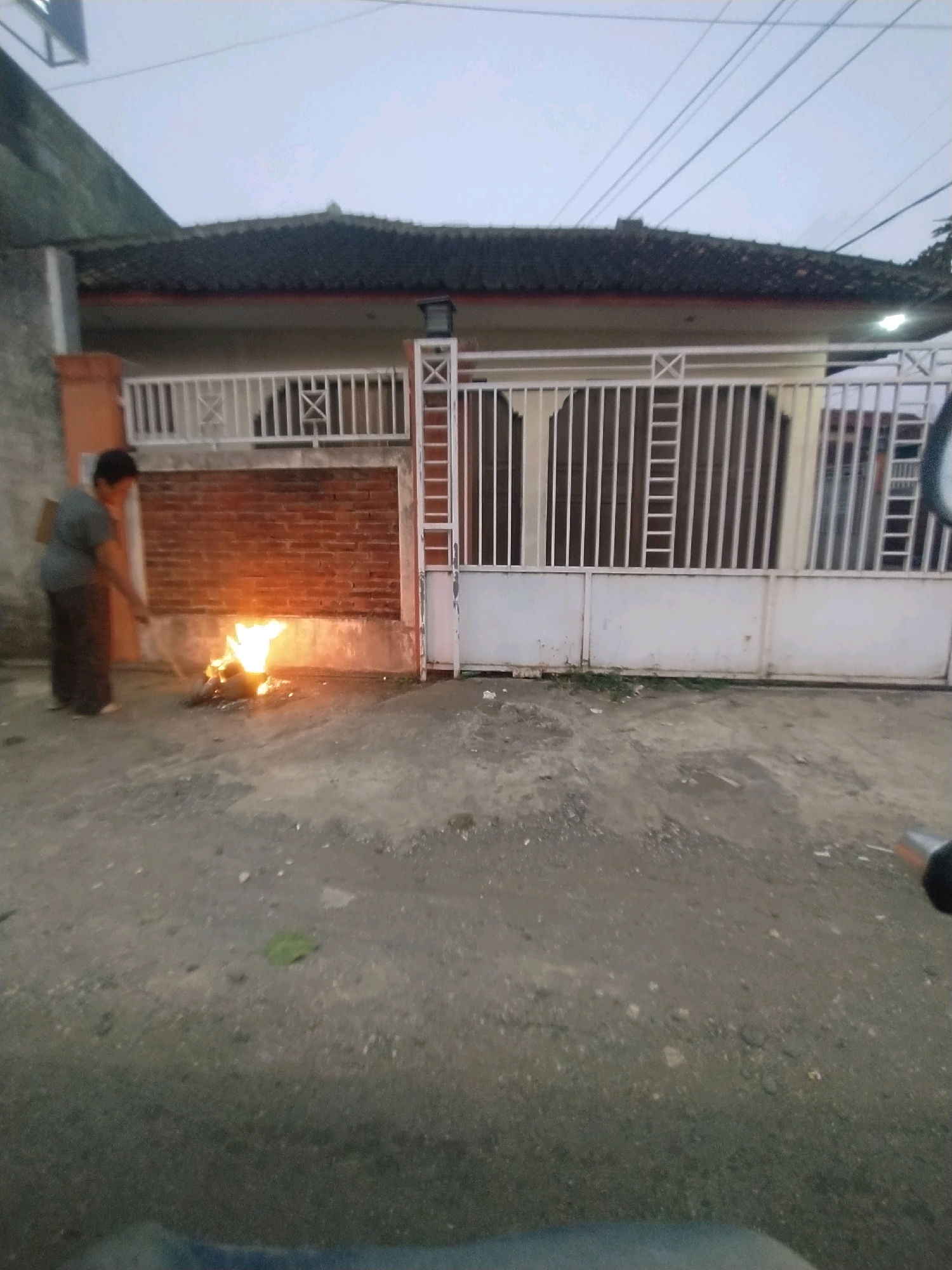 In Desa Karangbenda, Indonesia, an individual wearing dark pants and a gray t-shirt is standing to the left of a brick wall, tending to a small, active fire on the ground. The fire is burning debris, producing visible flames and smoke. A white metal gate with vertical bars is visible in front of a building with a dark roof. The ground is a mixture of asphalt and gravel. The sky is overcast, suggesting early morning or late evening.