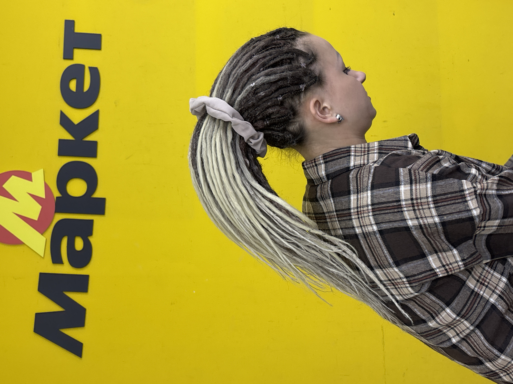 A woman with blonde dreadlocks tied in a ponytail stands in front of a yellow wall with the word "Market" and a stylized "M" logo. The photo was taken in the city of Chaikovsky, Russia. The woman is wearing a brown and white plaid shirt and a small earring. She is facing away from the camera and looking up.