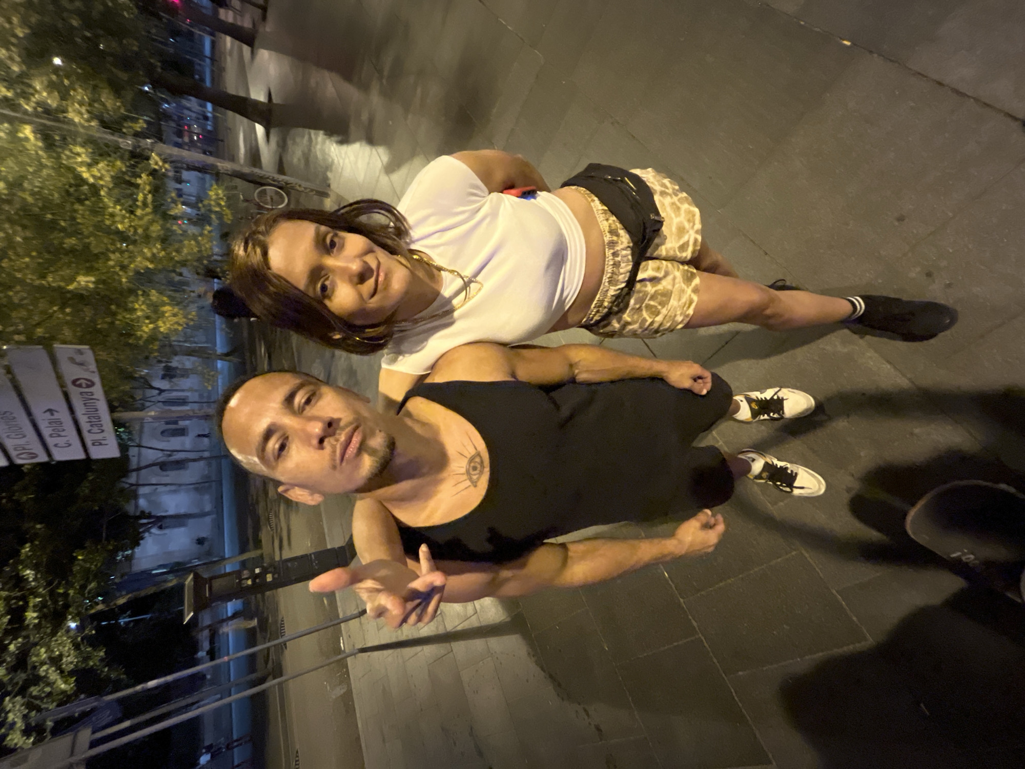 A low-angle, wide shot captures two individuals, a man and a woman, standing on a paved sidewalk at night. The man, on the left, is wearing a black tank top and white sneakers with black laces. He has a tattoo of an eye on his chest and is looking at the camera with a slight smirk. His left arm is around the woman's shoulders. The woman, on the right, is wearing a white crop top, patterned shorts with a black fanny pack around her waist, and black socks with white stripes tucked into black boots. She is smiling at the camera. Behind them, a street sign indicates directions to "Pl. Glòries," "C. Pelai," and "Pl. Catalunya," suggesting they are in Barcelona, Spain. Trees with yellow lights illuminating their leaves line the street, and the ground is wet, reflecting the light. The overall atmosphere is casual and social, possibly taken during a night out.