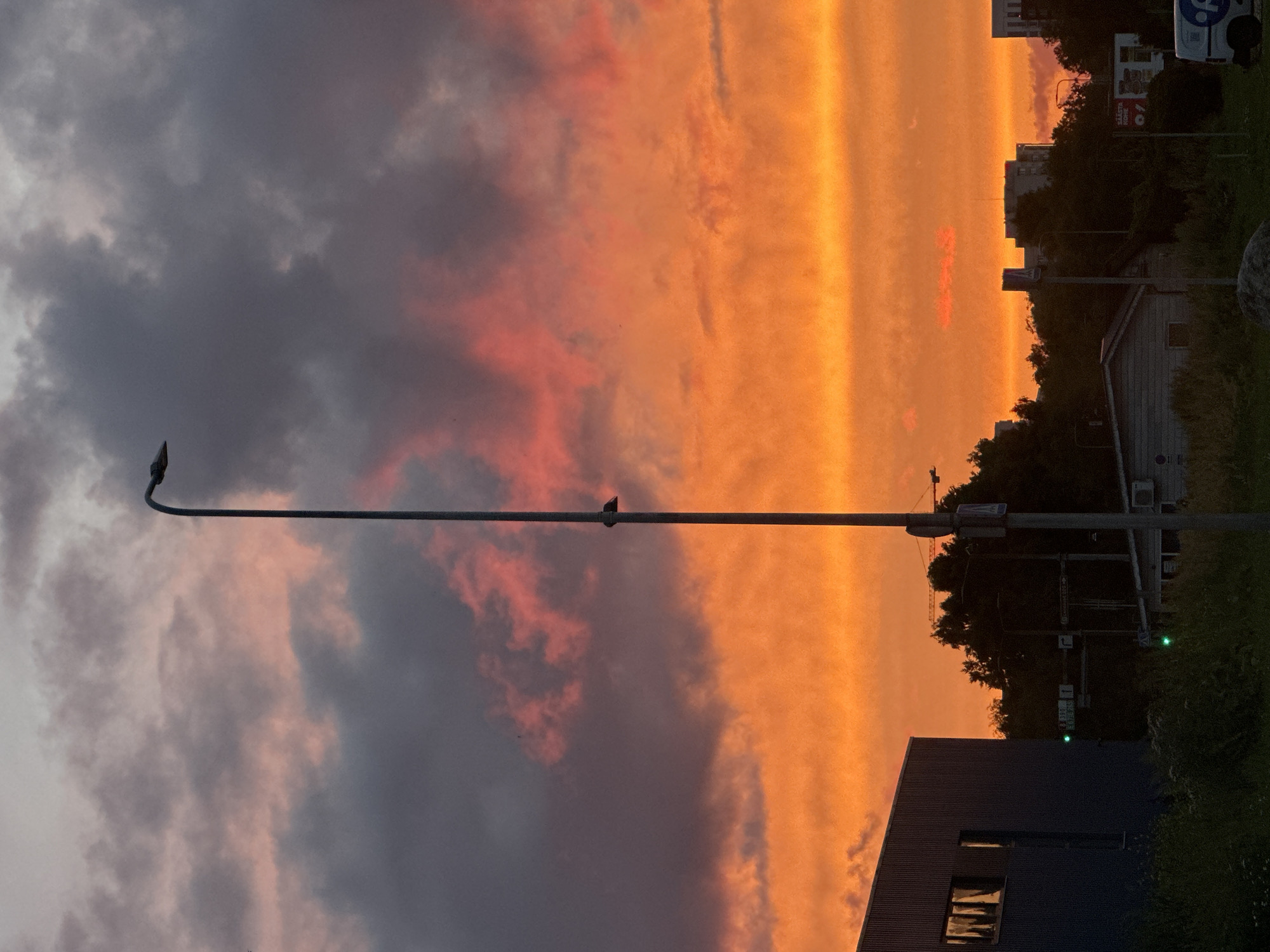 The image depicts a dramatic sunset over a cityscape, likely in Tallinn, Estonia, judging by the presence of a visible street sign with Cyrillic or Latin characters and the architectural style of the buildings. The sky is ablaze with vibrant orange and red hues, with streaks of pink and purple in the darker, upper clouds. A tall, slender lamppost stands prominently in the foreground, casting a long shadow. To the left, a dark blue, industrial-style building with reflective windows is visible. In the middle ground, silhouetted trees obscure a few buildings, and traffic lights show green signals. A construction crane can be seen in the distance, suggesting ongoing development. The lower part of the image shows a grassy area with some rocks, hinting at a park or undeveloped land. The overall scene captures the beauty of twilight in an urban setting.
