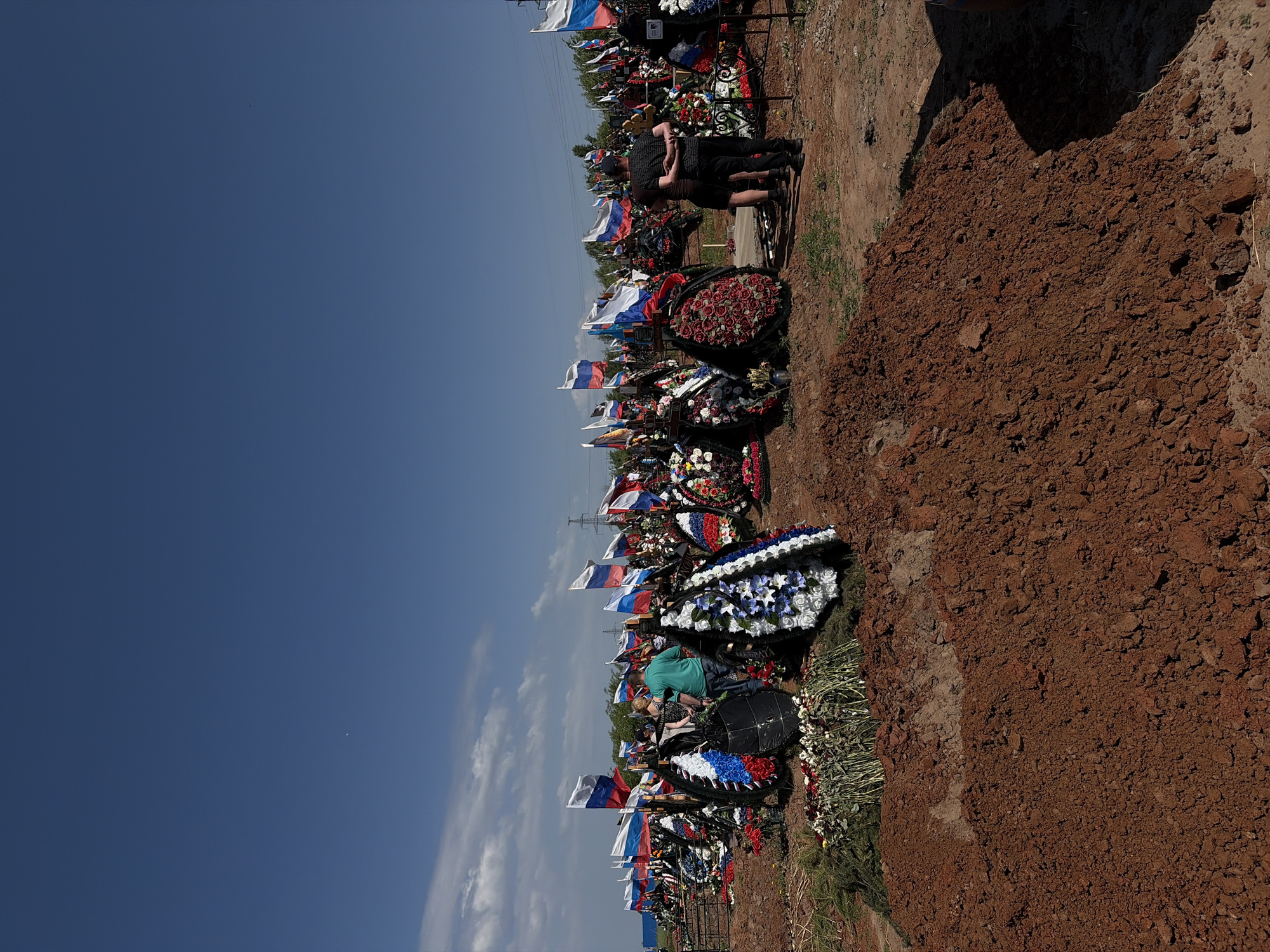 The image shows a cemetery in Izhevsk, Russia, with numerous graves adorned with wreaths and Russian flags. People are gathered among the graves, seemingly paying their respects. The ground is covered in red-brown earth. The sky is blue with scattered clouds.