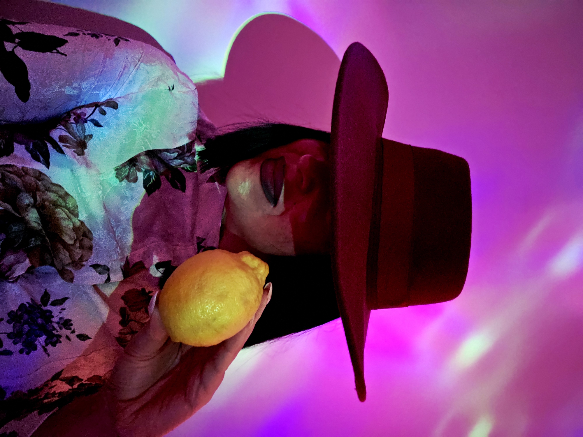 The vertically oriented image captures a person in an abstract, artificially lit environment. The subject, seen from the upper torso, wears a wide-brimmed, dark reddish-brown hat and a flowing garment with dark floral patterns against a backdrop of swirling purples, blues, and hints of green. The person's face is partially visible, with dark hair, and they are sticking their tongue out, suggesting a playful or sour expression. One hand, adorned with light nail polish, prominently holds a bright yellow lemon. The background is a vibrant wash of strong pinks, purples, and blues, featuring diffused light patterns that indicate dynamic, colored lighting. The atmosphere is surreal and artistic due to the striking color palette and lighting. There is no visible text, and the time of day or specific location is obscured by the artificial illumination.
