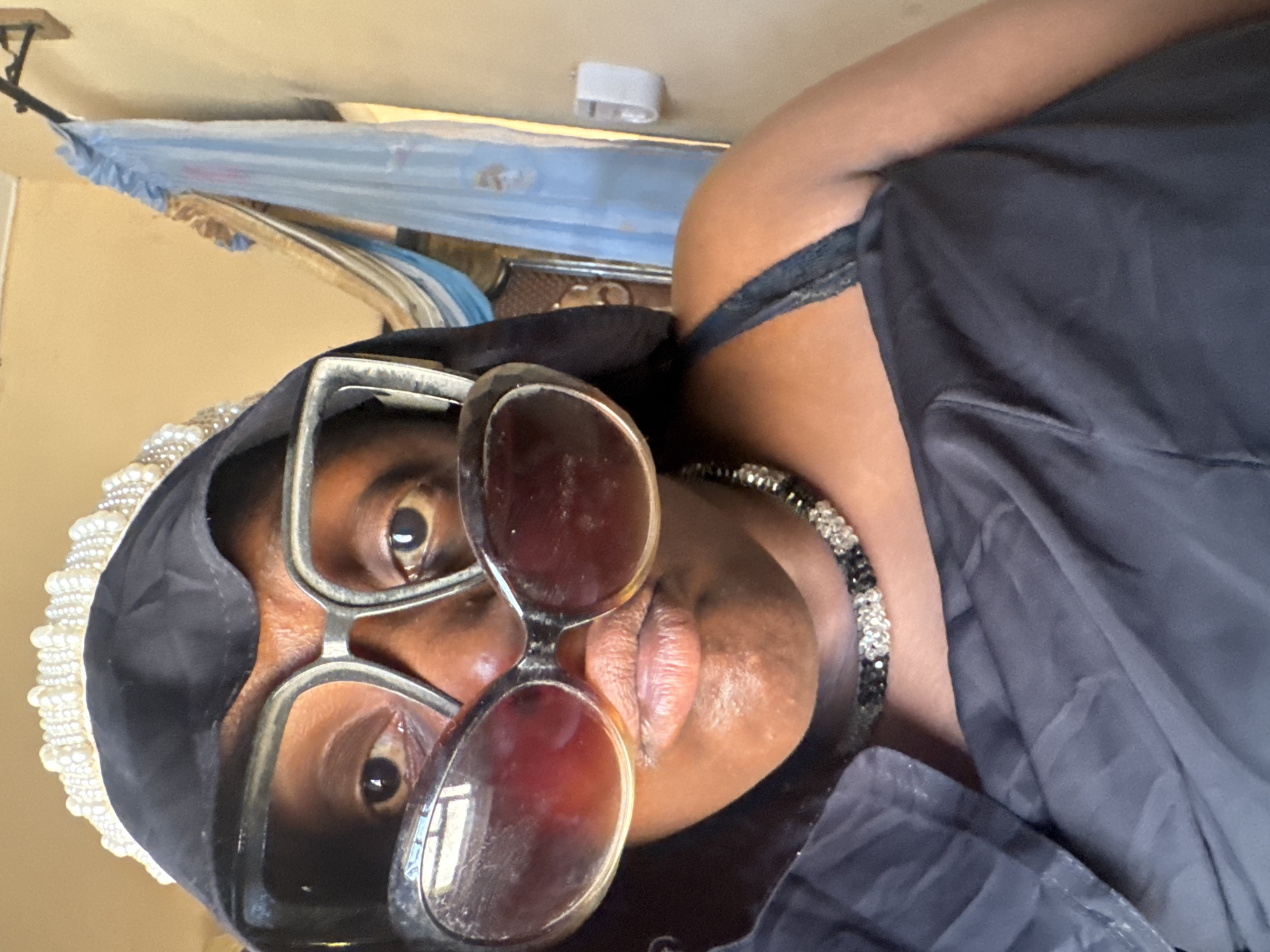 A person with dark skin is wearing oversized sunglasses with rounded frames, a black headscarf with white beaded embellishments along the edge, and a black top. They are also wearing a black and silver beaded necklace. The background includes a light blue fabric hanging from a rod, suggesting an interior space. The location is unknown; no information suggests this is Madangala, Nigeria.