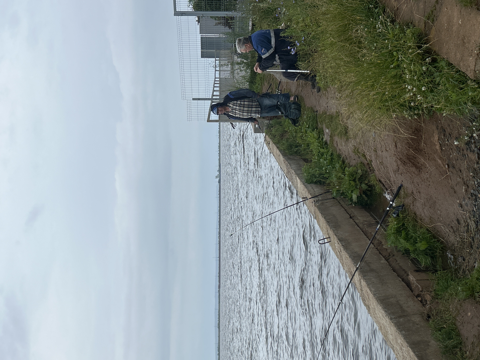 Two men are fishing on the shore of the Kama River in Chaikovsky, Russia. The weather is overcast. The men are standing and sitting on a concrete embankment. One man, wearing a plaid shirt and jeans, stands and holds a fishing rod. Beside him is a dark backpack. The other man, wearing a blue jacket, is seated on the ground and leans on a walking stick, also with a fishing rod. Fishing rods are also placed along the embankment, indicating active fishing. The river is choppy with waves. In the background, a wire fence encloses an industrial or utility area. The shore is overgrown with grass and some small wildflowers.