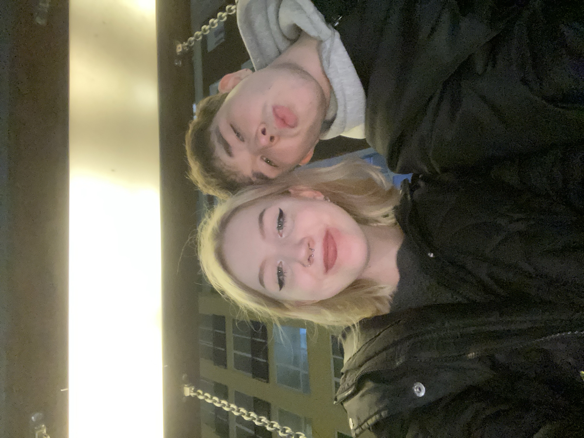 Two people are posing for a selfie. A light-skinned person with blond hair and a nose ring is in the foreground. Behind and slightly above them, another light-skinned person with brown hair is sticking their tongue out. Both are wearing dark-colored jackets. A building with illuminated windows is visible in the background. A chain is visible on the left side of the image. The location cannot be determined without additional context.