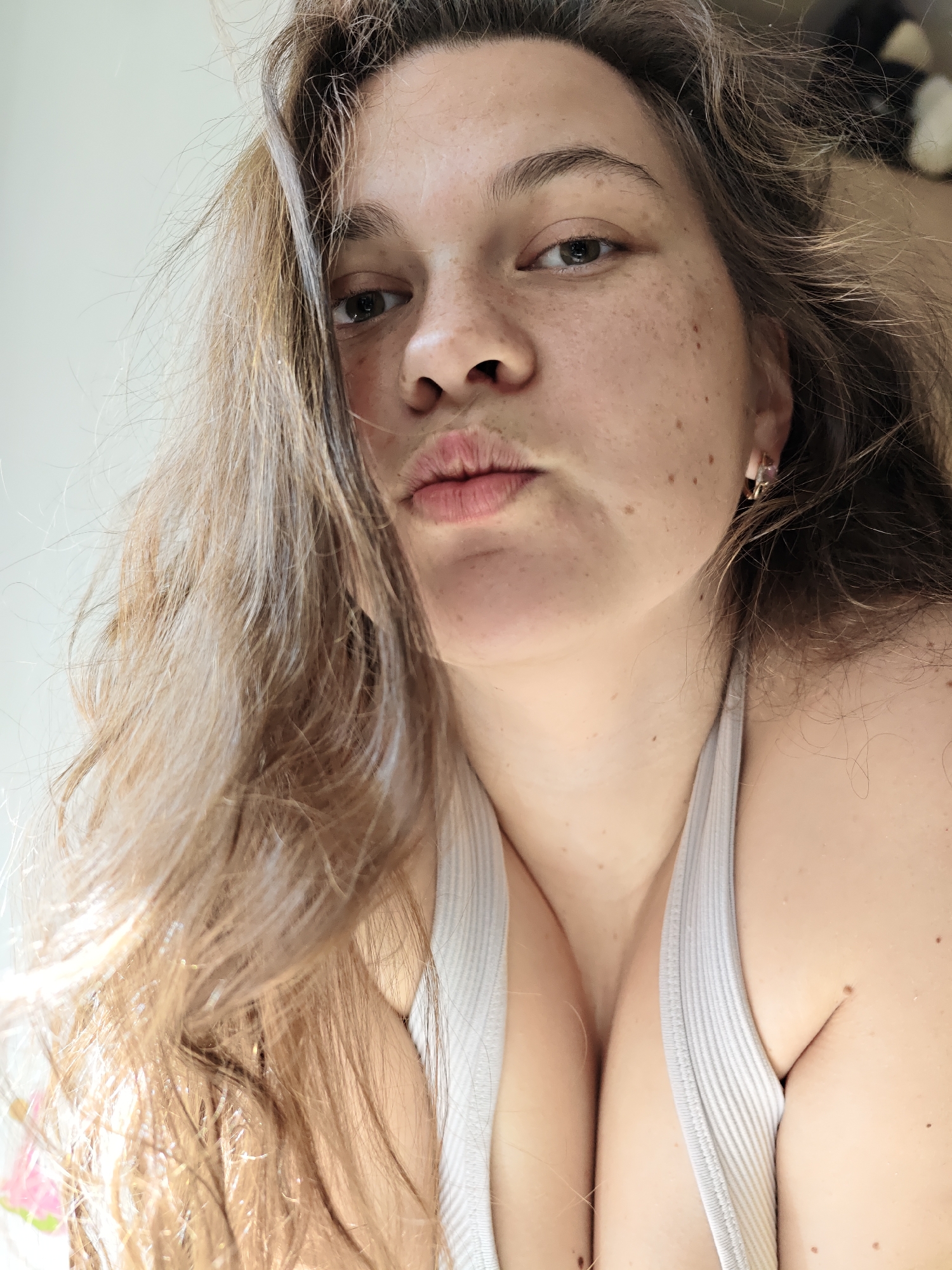 A woman with long, light brown hair and freckles is looking directly at the camera with pursed lips. She is wearing a light-colored ribbed tank top. A portion of her chest is visible. The background is a mix of light colors, suggesting an indoor setting. There is no indication of the location being in the city of Chaikovsky, Russia, or any specific city for that matter.
