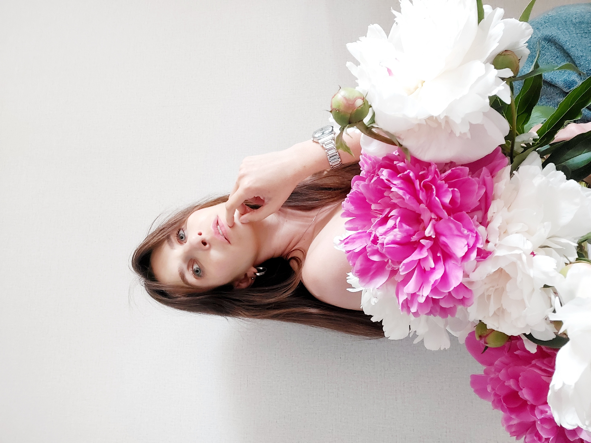 The image features a young woman posing amidst a large bouquet of peonies, set against a plain, light-colored background. The photo is oriented with the woman's head towards the bottom left, lying down with her gaze directed towards the viewer.
**Subjects:** The primary subjects are a young woman and a substantial arrangement of blooming peonies.
**Activity or event taking place:** The woman is engaged in a posed portrait, likely taken from an overhead perspective while she is reclining. Her left hand is gently placed near her chin or lower lip.
**Notable details:**
* **The Woman:** She has long, dark brown hair that spreads out around her head. Her eyes are light-colored, possibly blue or grey, and her expression is serene and direct, engaging with the camera. She wears a silver-toned wristwatch with a metal band on her left wrist and a simple silver hoop earring in her visible right ear. Her left shoulder is bare, suggesting a strapless garment or partial nudity, with the rest of her body mostly obscured by the flowers or out of frame.
* **The Peonies:** The bouquet is abundant, featuring large, full blooms of peonies in two prominent colors: vibrant fuchsia pink and pure white. Several green buds and leaves are interspersed among the open flowers, indicating freshness.
* **Setting and Background:** The background is an uncluttered, plain light wall, likely white or a very pale grey, providing a minimalist context that highlights the subjects. A small section of blue denim fabric is visible in the upper right portion of the image, behind the flowers, possibly part of the woman's attire (e.g., jeans) or an adjacent object.
* **Lighting:** The scene is illuminated by soft, diffused light, which casts subtle shadows and enhances the textures of the flowers and the woman's skin.
* **Location Cues:** None explicitly visible within the image, but the provided context states Chaikovsky, Russia.
* **Emotions:** The woman's calm gaze and gentle pose convey a tranquil and contemplative mood.
**Visible text:** There is no visible text in the media file.