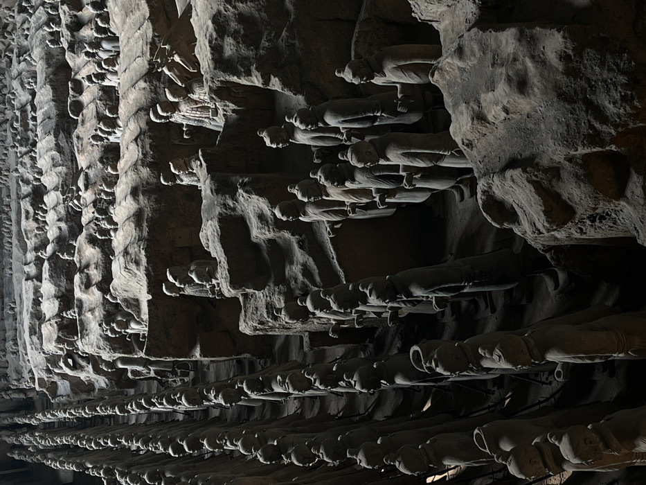 The image depicts a vast subterranean excavation pit at the Mausoleum of the First Qin Emperor in Lintong District, China. Thousands of life-sized terracotta warrior and horse statues are meticulously arranged in military formations within earthen trenches and along uneven earthen platforms. The foreground features multiple rows of standing warrior figures, primarily seen from the side or slightly angled, facing towards the left. These figures exhibit distinct uniforms, headwear, and facial features, predominantly in varying shades of grey and brown terracotta. Some statues are fully reconstructed and stand upright, while others in the background and midground are partially obscured by excavated earth, indicating various stages of preservation or ongoing archaeological work. The environment consists of rough, textured earthen walls and ground, with artificial lighting creating shadows and highlighting the sculptural details and the layered structure of the pit. No live individuals are present in the scene. The figures are static, depicting an ancient imperial army in perpetual formation.