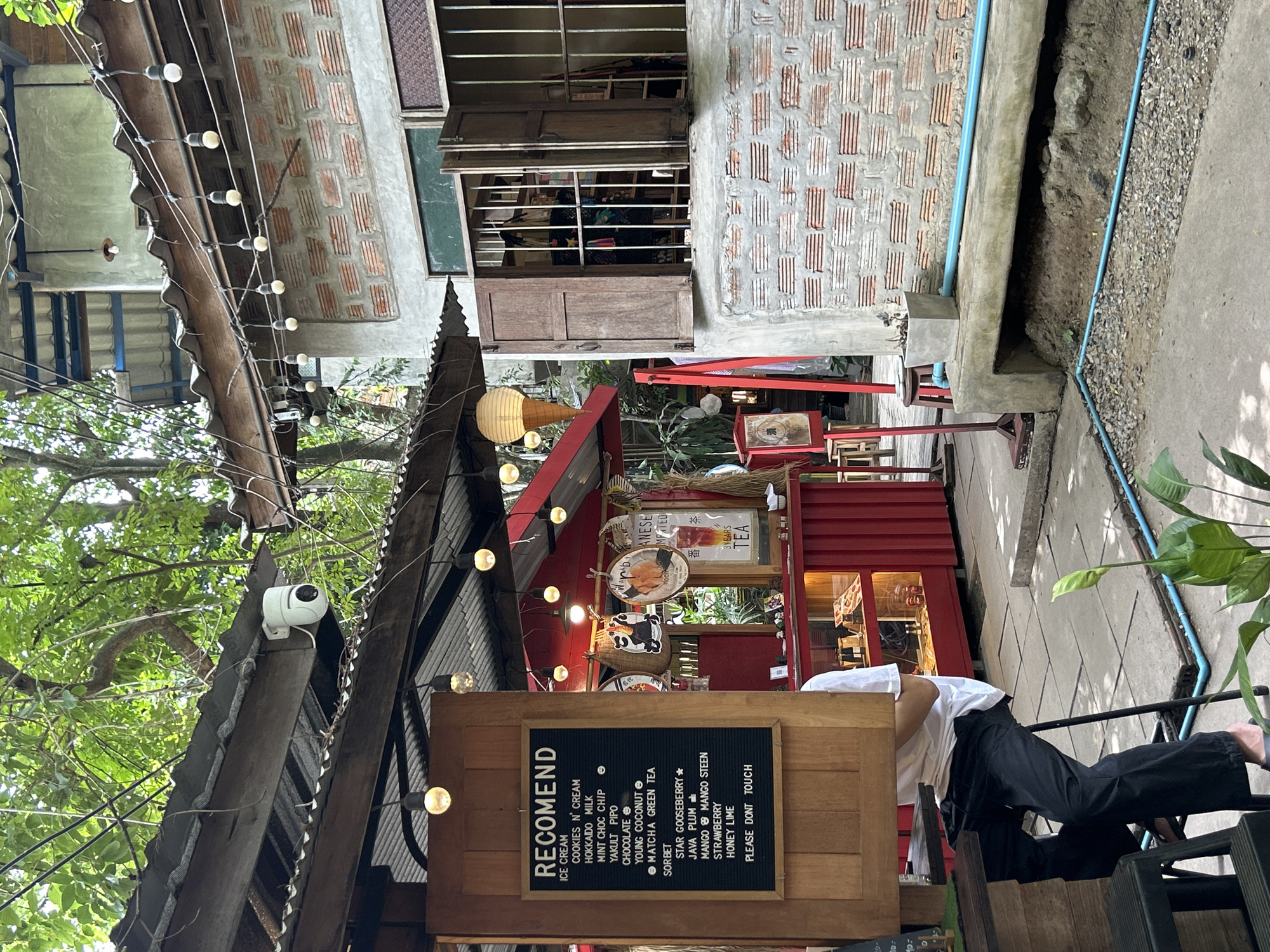 The image depicts an outdoor seating area at what appears to be a small cafe or food stall in Ban Sai Kham, Thailand. In the foreground, a person is seated on a high stool, wearing a white t-shirt and dark trousers, partially obscured by a wooden structure. A menu board to the left lists various ice cream and sorbet flavors, including "Cookies N' Cream," "Hokkaido Milk," "Mint Choc Chip," "Yakult Pipo," "Chocolate," "Young Coconut," and "Matcha Green Tea," as well as sorbets like "Star Gooseberry," "Java Plum," "Mango Mango Steen," "Strawberry," and "Honey Lime."
Behind the seated individual, a red stall with glass display cases is visible, showcasing various items, possibly food or merchandise. Decorative elements, including paper lanterns shaped like ice cream cones and other hanging ornaments, are present. A brick building with wooden shutters and barred windows stands to the right of the stall. The exterior of this building is constructed with rough concrete and exposed brickwork. Outdoor lighting, in the form of string lights with bare bulbs, hangs from a corrugated metal roof structure. The ground is paved with concrete slabs, with some gravel and exposed soil visible. A blue pipe runs along the ground and the base of the concrete structure. Lush green foliage from trees is visible overhead, indicating an outdoor, possibly tropical, setting. A white security camera is mounted on the underside of the roof.