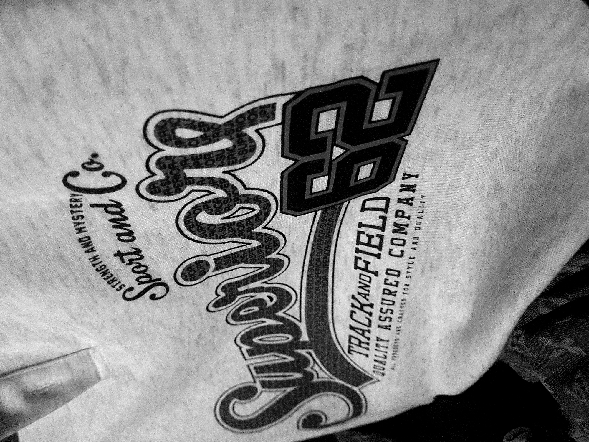 A grayscale, close-up shot shows a light-colored t-shirt with a graphic design. The design features bold, stylized text. At the top, "STRENGTH AND MYSTERY" is written in a smaller, serif font. Below this, "Sport and Co." is written in a large, flowing script font. The word "Super" is emblazoned across the center of the shirt in a thick, shadowed, curved font, with "IOR" repeated within the letters. To the right of "Super," the number "62" is prominently displayed in a blocky, outlined font. Beneath "Super 62," the text "TRACK AND FIELD" is printed in a capital serif font, followed by "QUALITY ASSURED COMPANY" in a smaller serif font. At the very bottom, a line of smaller text reads, "ALL PRODUCTS ARE CRAFTED FOR STYLE AND QUALITY." The t-shirt appears to be made of a textured fabric, with a subtle ribbed collar visible at the top left. The background is blurred and dark, with some textured fabric appearing in the lower left corner, suggesting it may be a person wearing the t-shirt. The location is Unknown, Unknown.