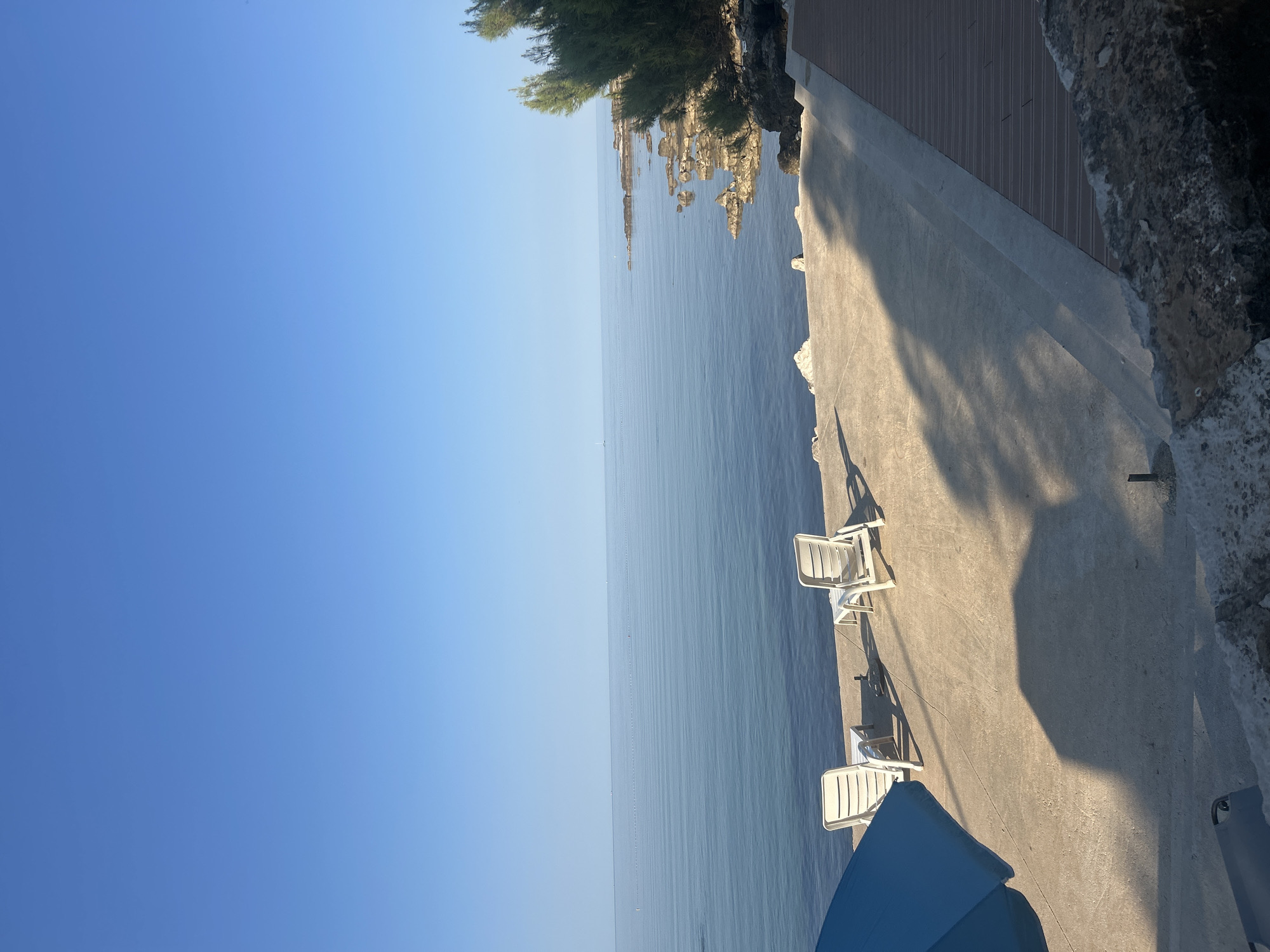 In Katoro, Croatia, a tranquil scene unfolds by the sea. The image captures a calm, blue ocean under a clear sky. In the foreground, two white lounge chairs are set on a concrete patio, positioned to face the water. To the left, a portion of a blue umbrella is visible, casting a shadow on the patio. The patio itself is bordered by a rocky shoreline with sparse green vegetation. A wooden deck runs along the right side of the patio. The overall atmosphere is one of peaceful relaxation and natural beauty.