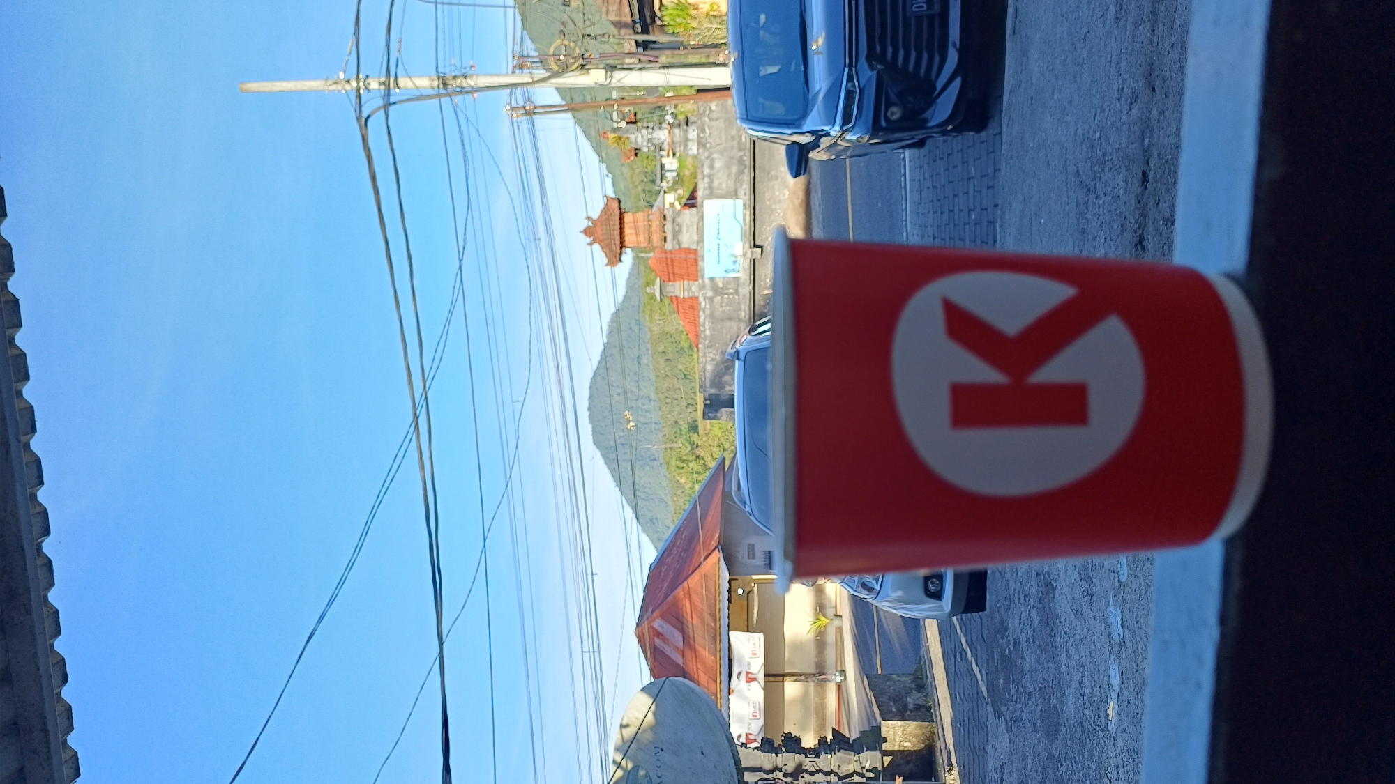 Here is a detailed description of the image:
**Scene:** A roadside view in Baturiti, Indonesia, oriented vertically. The prominent foreground feature is a red paper cup with the white "K" logo of Circle K convenience store. The cup rests on a ledge, potentially a wall or a window frame.
**Environment:** The background shows a street scene on a sunny day. A mountain covered in greenery looms in the distance, with a traditional Balinese gate-like structure visible in front of it. Utility poles and wires stretch across a bright blue sky.
**Objects:** Several vehicles are parked on the roadside, including a dark blue car and a white vehicle. A store front is visible.