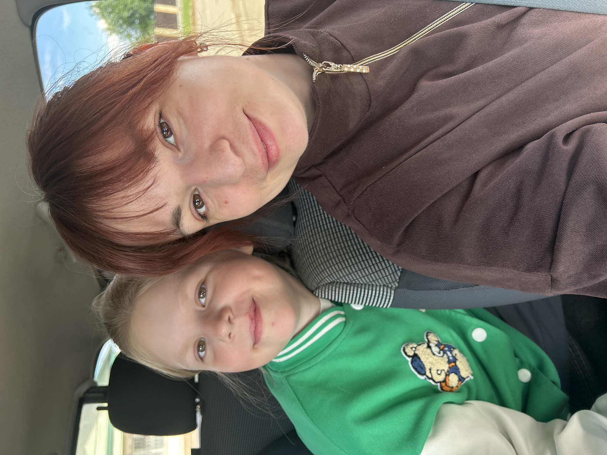 The image depicts a selfie taken inside a car, showing an adult female and a young child. The woman has reddish-brown hair and is wearing a brown sweater with a gold zipper. The child, who appears to be a girl, has blonde hair and is wearing a bright green jacket with white stripes and a cartoon character patch. The seatbelts are visible, suggesting they are riding in the car. The environment outside the car is a building and trees, indicating an urban setting. There is no information in the image confirming it is Фоки, Russia.