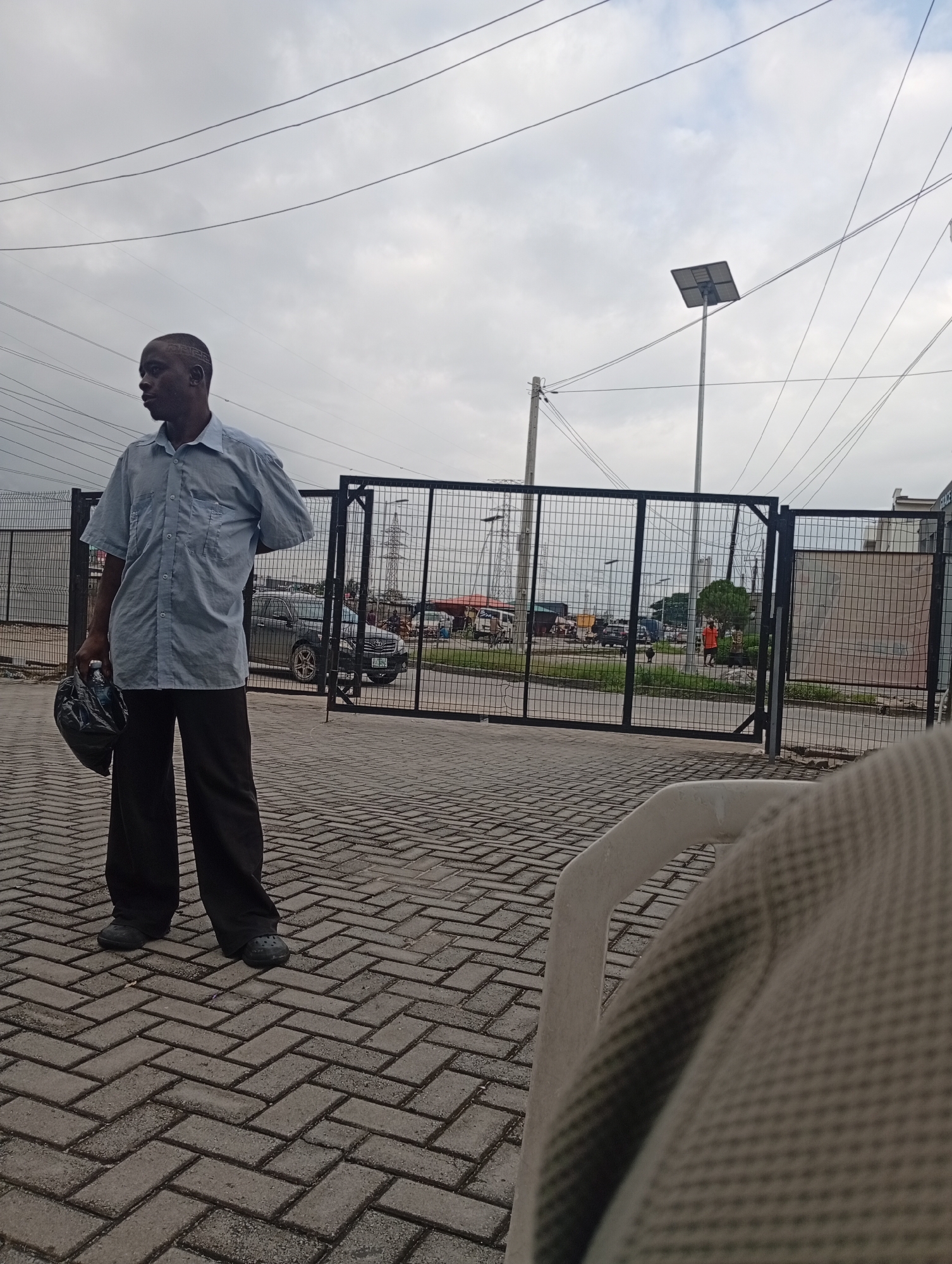 A man stands facing left on a brick-paved walkway outside a fenced area, holding a black bag in his left hand. He is wearing a light blue short-sleeved collared shirt and dark pants. In the background, through the black metal fence, there are several vehicles, including a dark sedan, and a person wearing an orange shirt. The environment appears to be outdoors with a cloudy sky. There are power lines visible overhead and a solar-powered street light. The paved area extends to the fence, and beyond the fence is a grassy area with some trees. The overall scene is located in the city of Ilado, Nigeria.