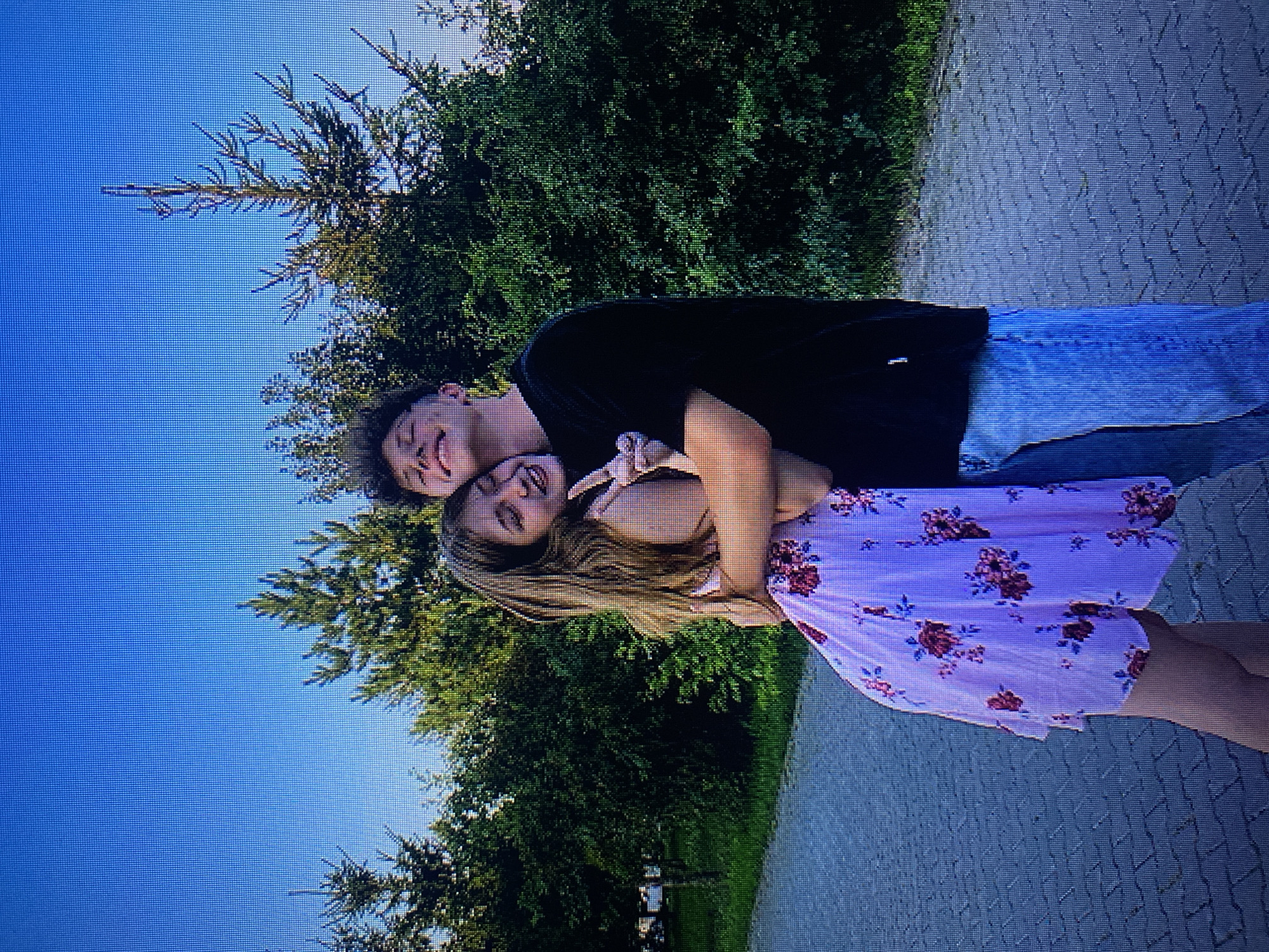A young couple is embracing in front of a line of trees under a clear blue sky. The female is on the left, wearing a light pink dress with a floral pattern. She has long, light brown hair and is smiling with her eyes closed. She is making a peace sign with her right hand. The male is on the right, wearing a black t-shirt and blue jeans. He has short, curly brown hair and is smiling with his eyes closed, leaning his head down to embrace her. They are standing on a paved walkway. The scene appears to be outdoors, possibly a park or a public area. There is no identifiable information in the image to suggest a specific location in Poznan, Poland.