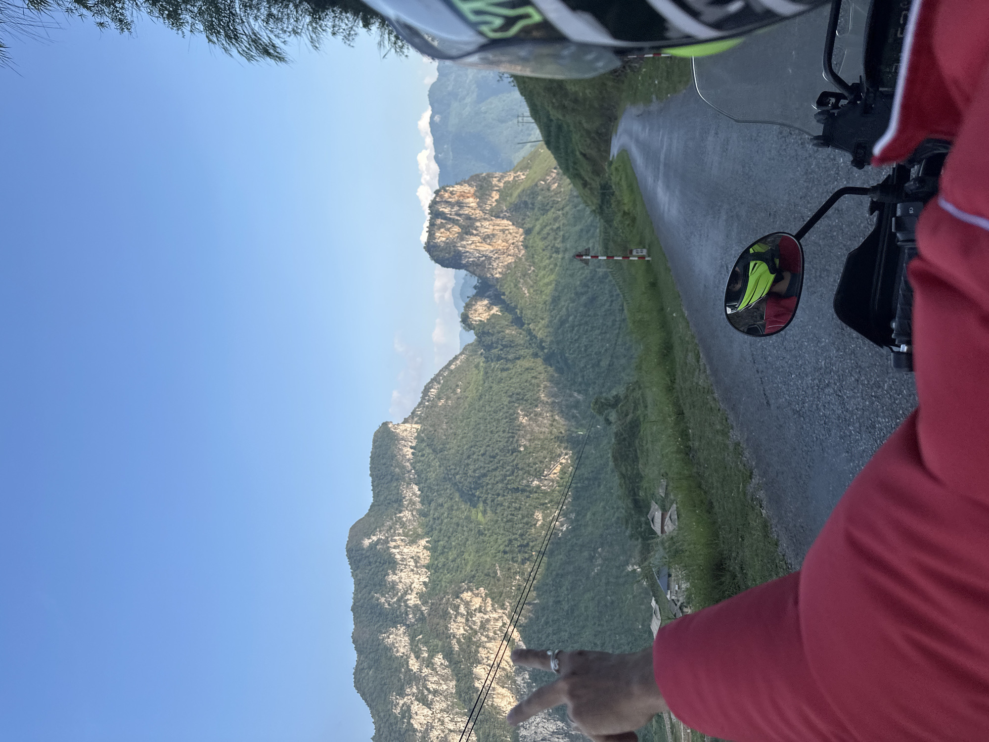 The image shows a first-person view from a motorcycle on a paved road in Đường Thượng Commune, Vietnam. The rider is wearing a helmet and a red jacket. In the motorcycle's side mirror, a person in a neon yellow vest can be seen. The road winds alongside a mountain range densely covered in green vegetation. A set of red and white striped guard posts are visible next to the road. The sky is clear and blue. A hand is pointing towards two power lines.