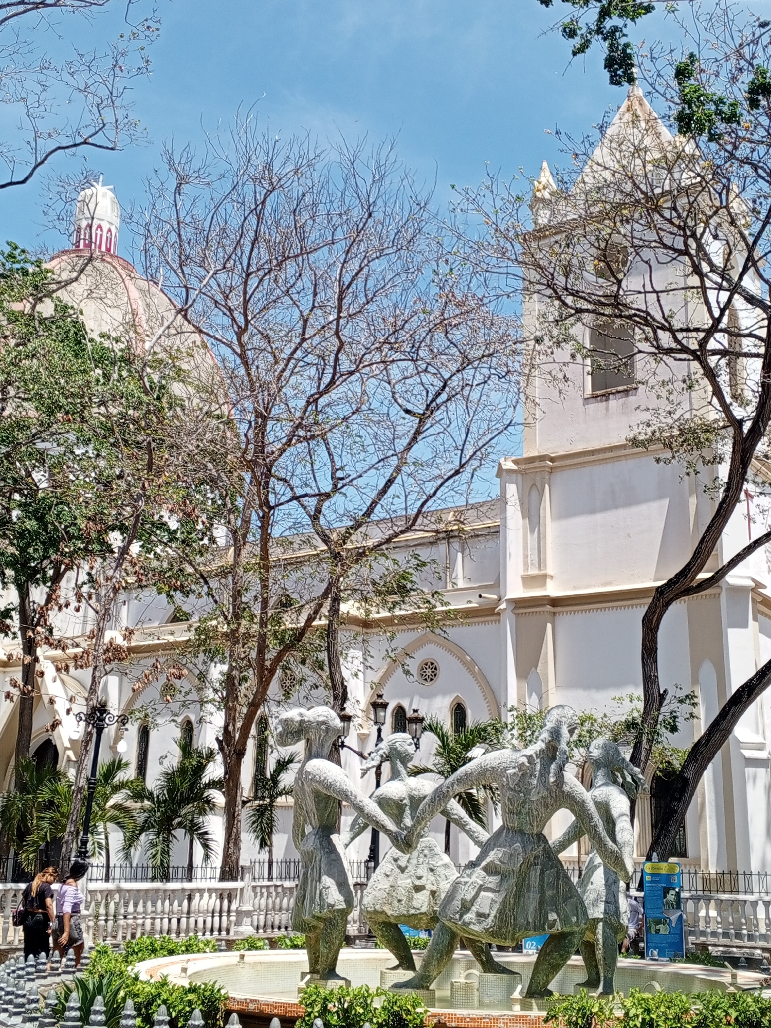 The image shows a sculpture of five dancing women in front of a large, white church. The church has a dome with a red and white cupola, and a tall, rectangular bell tower. Several trees with bare branches frame the buildings. A white, ornate fence surrounds the sculpture and fountain. In the background, a sign with information is visible to the right. In the distance, three women are walking along the sidewalk. The location is likely in Porlamar, Venezuela.