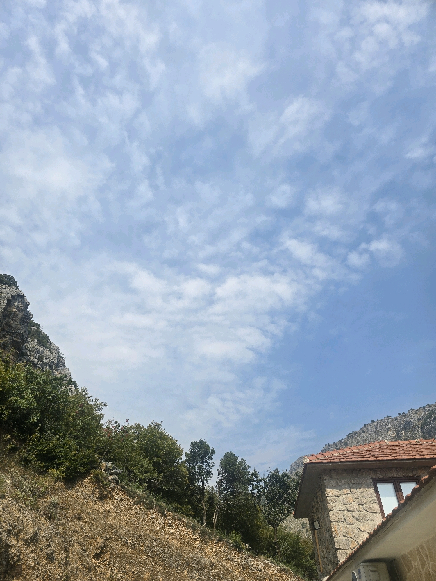 The image captures a mountainous landscape with a partial view of a stone building in the foreground. The sky is bright blue with scattered white clouds. To the left, a steep, rocky mountainside is covered with dense green trees and vegetation. The ground in the foreground is dry and rocky, with sparse grass. A portion of a building with a terracotta tiled roof and stone walls is visible on the right side of the image. A window with reflective glass is visible on the building. An air conditioning unit is mounted on the exterior wall of the building. The overall scene suggests a rural or semi-rural mountainous setting. The image is likely taken in a location similar to the mountainous regions surrounding Podgorica, Montenegro, known for their natural beauty and stone architecture. No people are visible in the image, and no specific actions or interactions are depicted.