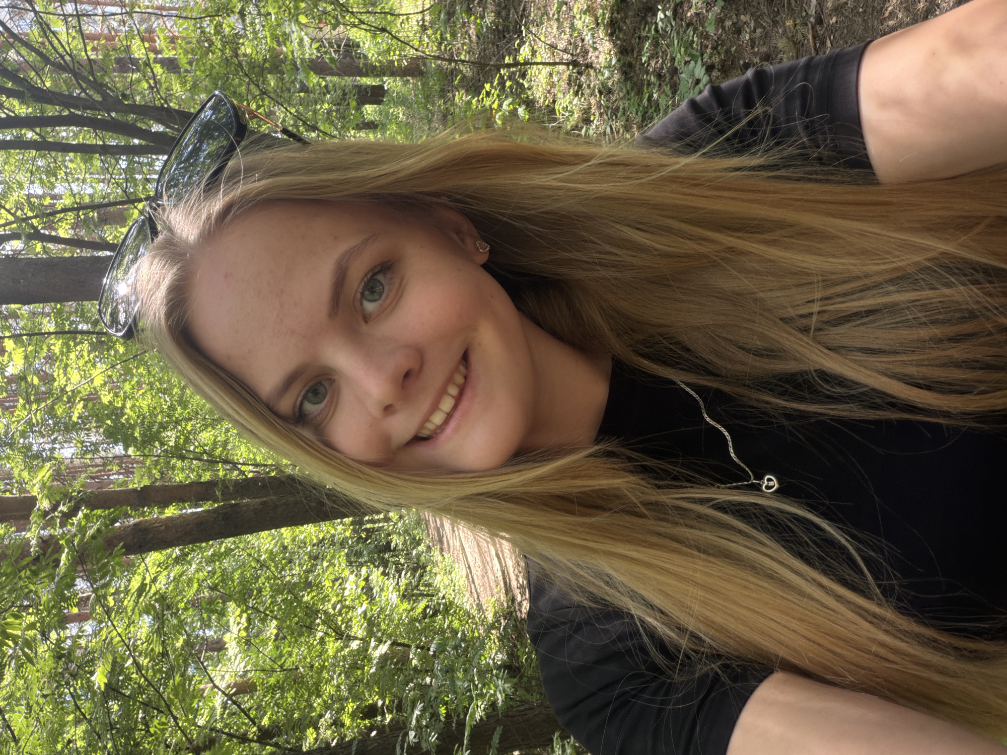 A woman with long, blonde hair is taking a selfie in a wooded area. She is wearing a black top and a silver necklace. Sunglasses are perched on her head. The background is out of focus, but it appears to be a forest with green trees and sunlight filtering through the leaves. The location in the city of Izhevsk, Russia is unknown.