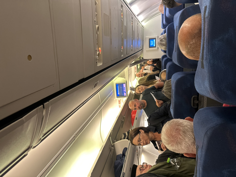 The image displays the interior of a commercial aircraft cabin, located in the city of Tzafria, Israel. The perspective is from a seated passenger, looking forward down a central aisle.
In the foreground, the backs of two dark blue upholstered airline seats are visible. The left seat partially reveals the head of a male passenger with short, grey hair, with a green-capped plastic water bottle visible near his left shoulder. The right seat shows the top of a male passenger's bald head, exposing reddish-brown hair on the sides.
The midground features a crowded aisle where multiple passengers are standing. A woman with dark hair and glasses, wearing a dark jacket, is prominent on the left, holding a small, dark rectangular object, possibly a boarding pass. To her right stands a bald man with a short beard, wearing a dark short-sleeved shirt, holding a white paper slip in his right hand. Behind them, additional passengers are visible, queuing in the aisle.
The aircraft cabin features white ceiling panels, integrated lighting, and overhead storage bins lining both sides. Some open sections of the left overhead bin contain blue bags. Two small, blue-lit LCD screens are mounted on the overhead panels. The screen on the left displays the text "Welcome Again" and "IAHOM", with "EXIT ESRE" visible below it. Another identical screen is visible further down the aisle on the right. The cabin is illuminated by artificial lighting. Rows of dark blue passenger seats, some occupied and some vacant, extend into the background.