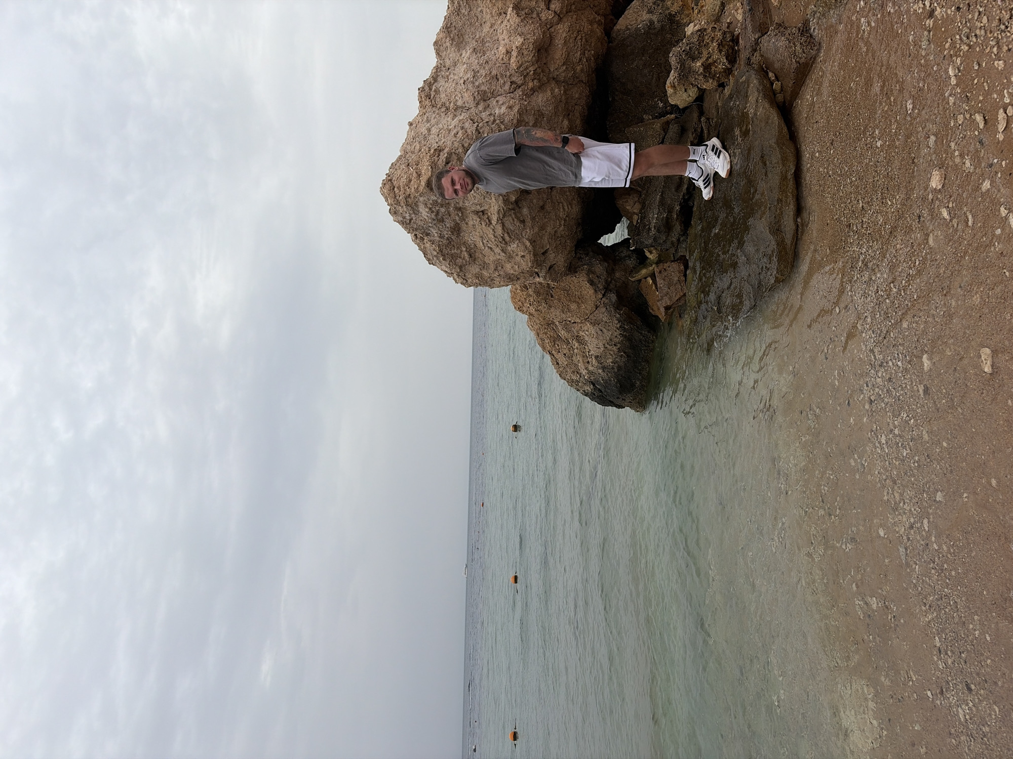 A man stands on a rocky outcrop at the edge of the sea. He is wearing a gray t-shirt, white shorts, and white sneakers with black stripes. The water is clear and extends to a line of buoys. The sky is overcast. This location appears to be in Sharm El Sheikh, Egypt.