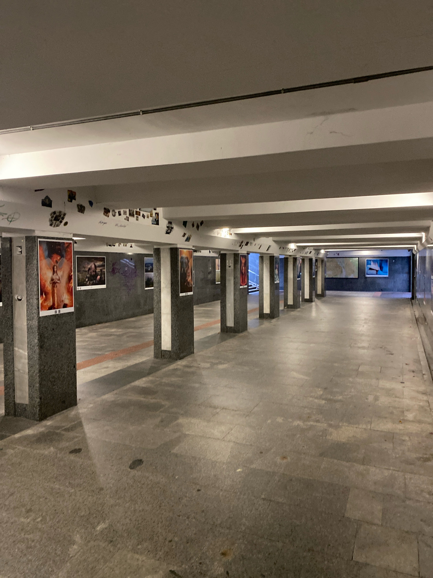 The image shows an underpass decorated with various artworks. The underpass is lined with granite-clad pillars that support the ceiling, from which hang framed pictures and smaller photographs. The walls are adorned with graffiti and additional framed images, including what appears to be a large map on the far wall. The floor is paved with large stone tiles, and a red line is visible on the ground, marking a pathway. The lighting is provided by fluorescent fixtures along the ceiling, casting shadows and illuminating the scene. The underpass is empty of people, creating a sense of quiet and stillness.
