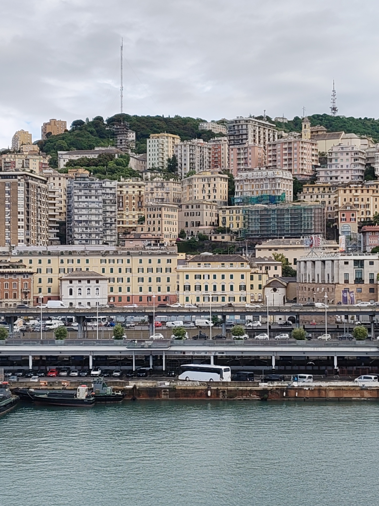 The image shows a cityscape of Genoa, Italy, with a harbor in the foreground. The water is calm. Several boats are docked at a concrete pier. Above the pier, there are two elevated roadways filled with cars and trees. Buildings of various heights, styles, and colors are densely packed on a hillside above the roadways. A tall communication tower is visible on the top of the hill. The sky is overcast.