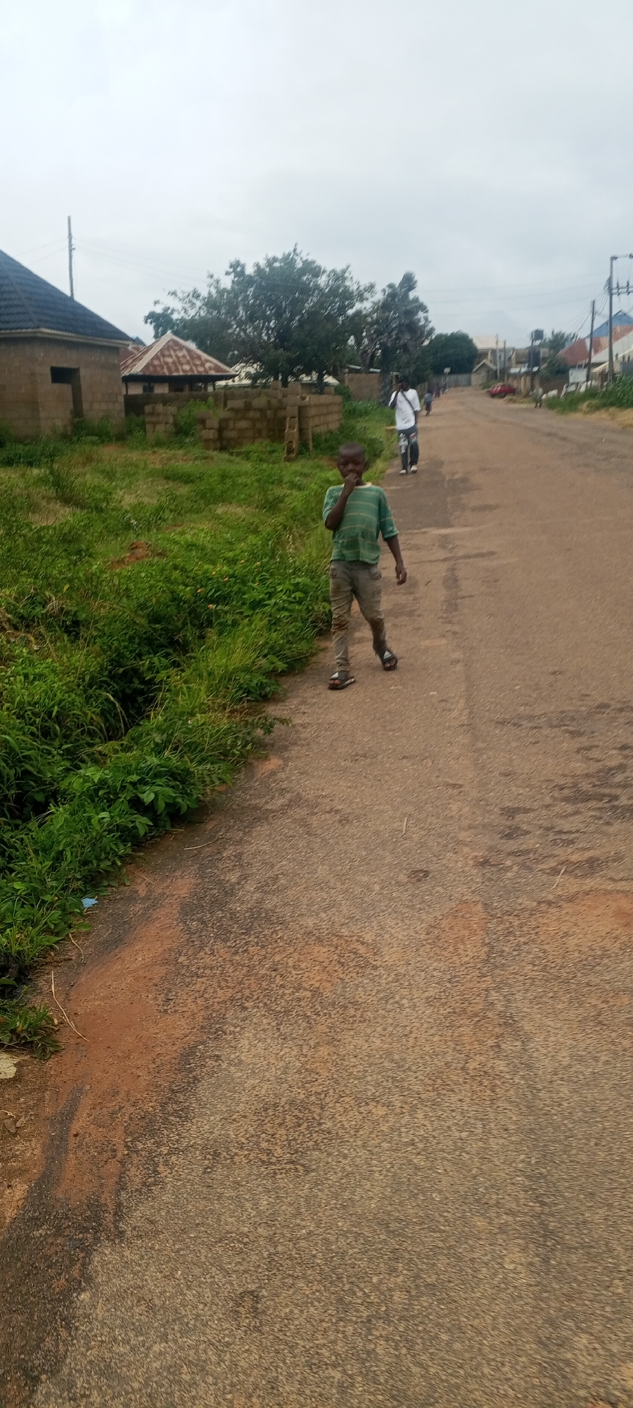 A young boy stands on an asphalt road in Bahit, Nigeria, his right hand near his mouth. He is wearing a horizontally striped green t-shirt and grey pants. He has sandals on his feet. To his left, there is a strip of green grass and a dirt embankment. Behind him, a man in a white shirt and dark pants walks away from the camera, and further down the road, there are buildings and trees. The sky is overcast.
