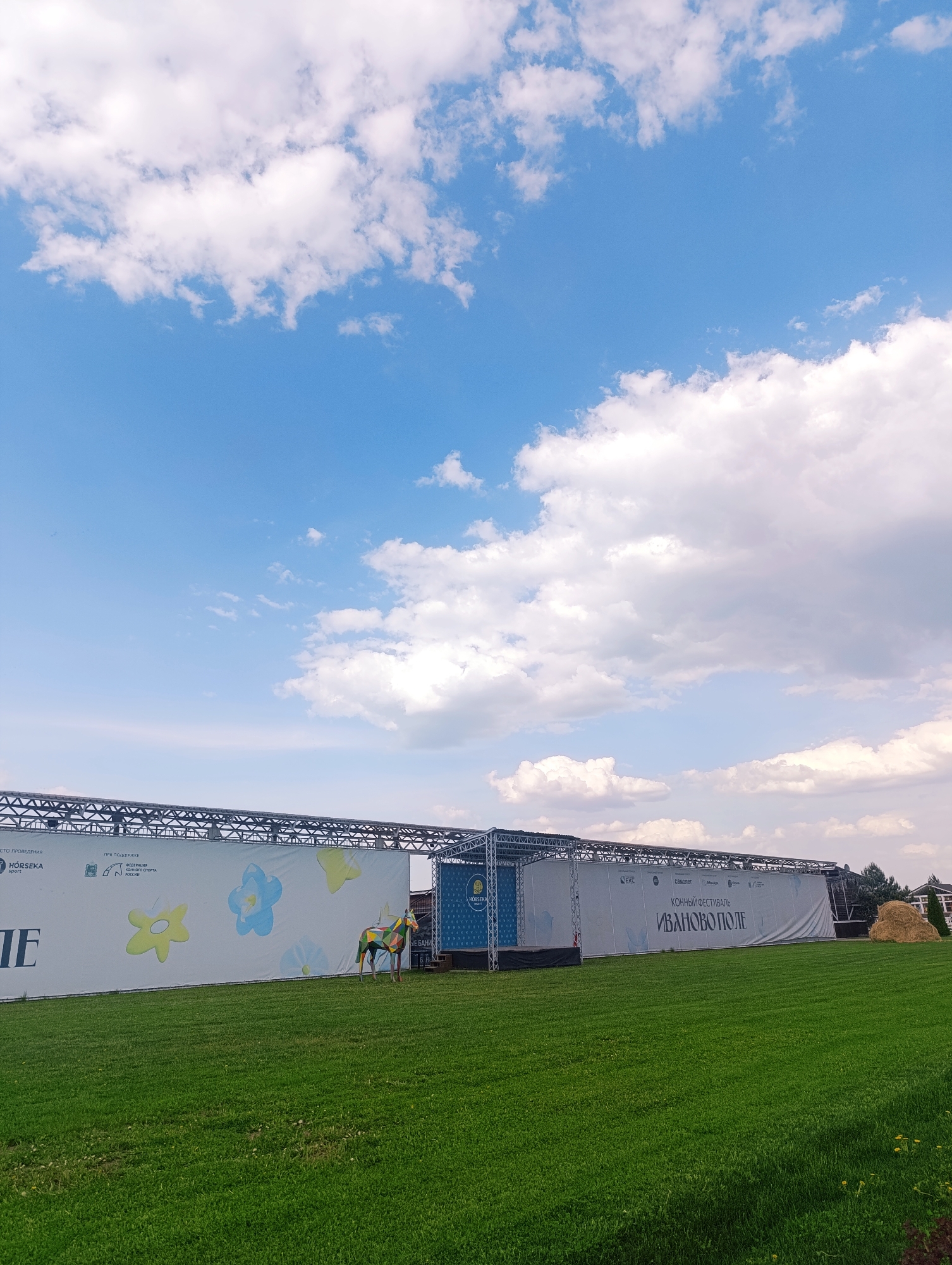 The image captures an outdoor scene under a blue sky dotted with fluffy white clouds. A large, rectangular structure stretches horizontally across the middle ground. The structure appears to be a temporary setup, likely for an event, as indicated by the banners. One banner displays the text "СТО ПРОВЕДЕНИЯ" and includes the word "HÓRSEKA sport." Another banner reads "ПРИ ПОДДЕРЖКЕ ФЕДЕРАЦИЯ КОННОГО СПОРТА РОССИИ." A third banner contains the text "HORSEKA" along with "EKC," "самолет," and "ИВАНОВО ПОЛЕ," also noting "КОННЫЙ ФЕСТИВАЛЬ" (Horse Festival). In front of the structure is a well-maintained green lawn. There is a life-sized, colorfully painted statue of a horse. In the background, a glimpse of trees and a hay bale, and what appears to be a traditional Russian wooden house can be seen on the right side. This scene is located in the city of Ivanovskoe, Russia, judging from the text on the banner.