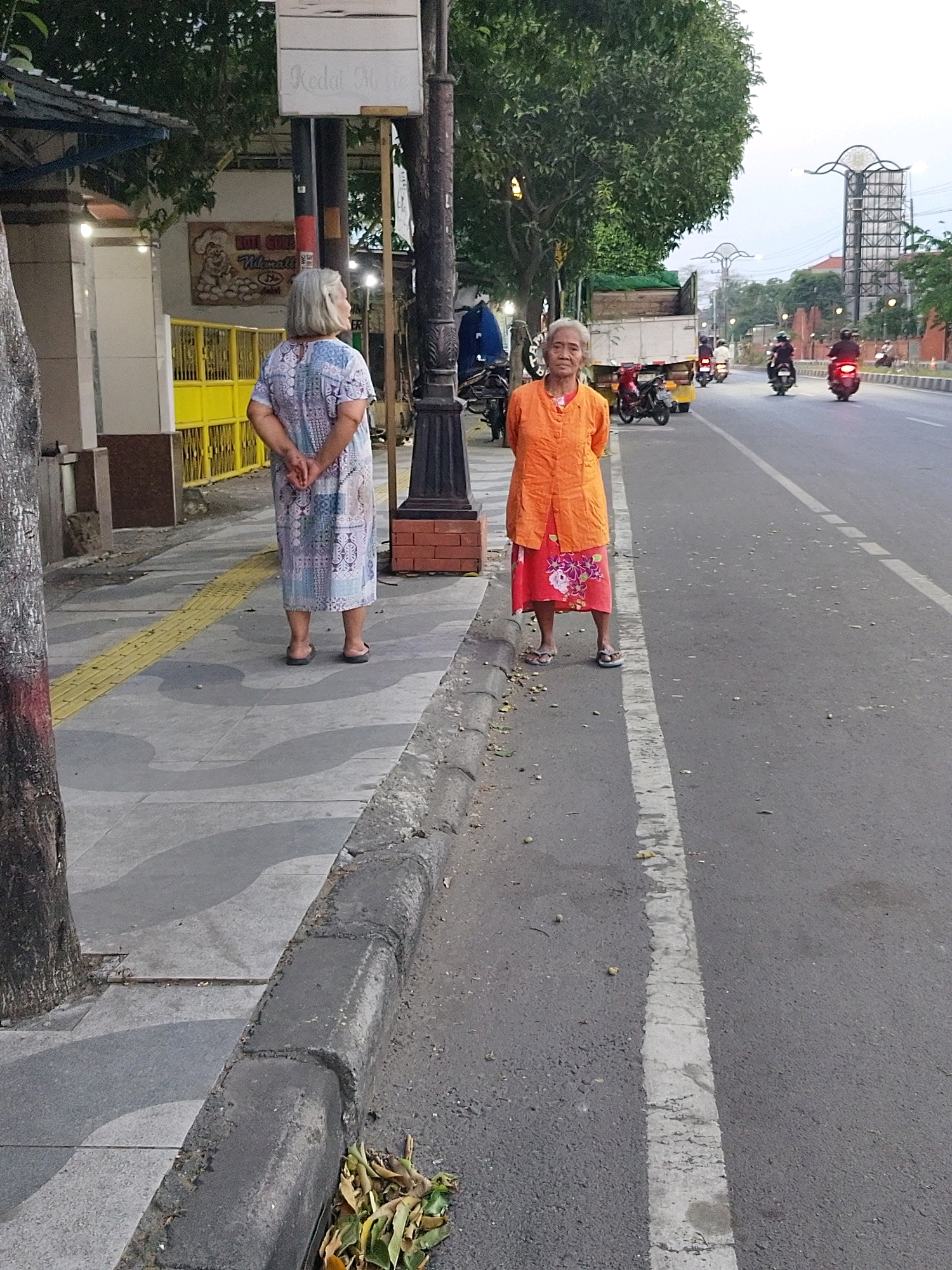 Two elderly Indonesian women are standing on a sidewalk next to a street in Mojokerto, Indonesia. The woman on the left is wearing a light blue and white patterned dress and appears to be facing away from the camera. The woman on the right is wearing an orange top and a floral-patterned skirt and is facing the camera. There are motorcycles and cars on the street. A street lamp is visible on the sidewalk behind the women. The sidewalk has a yellow tactile paving strip. The buildings on the left of the image are partially visible, and trees line the street. The sky is overcast.