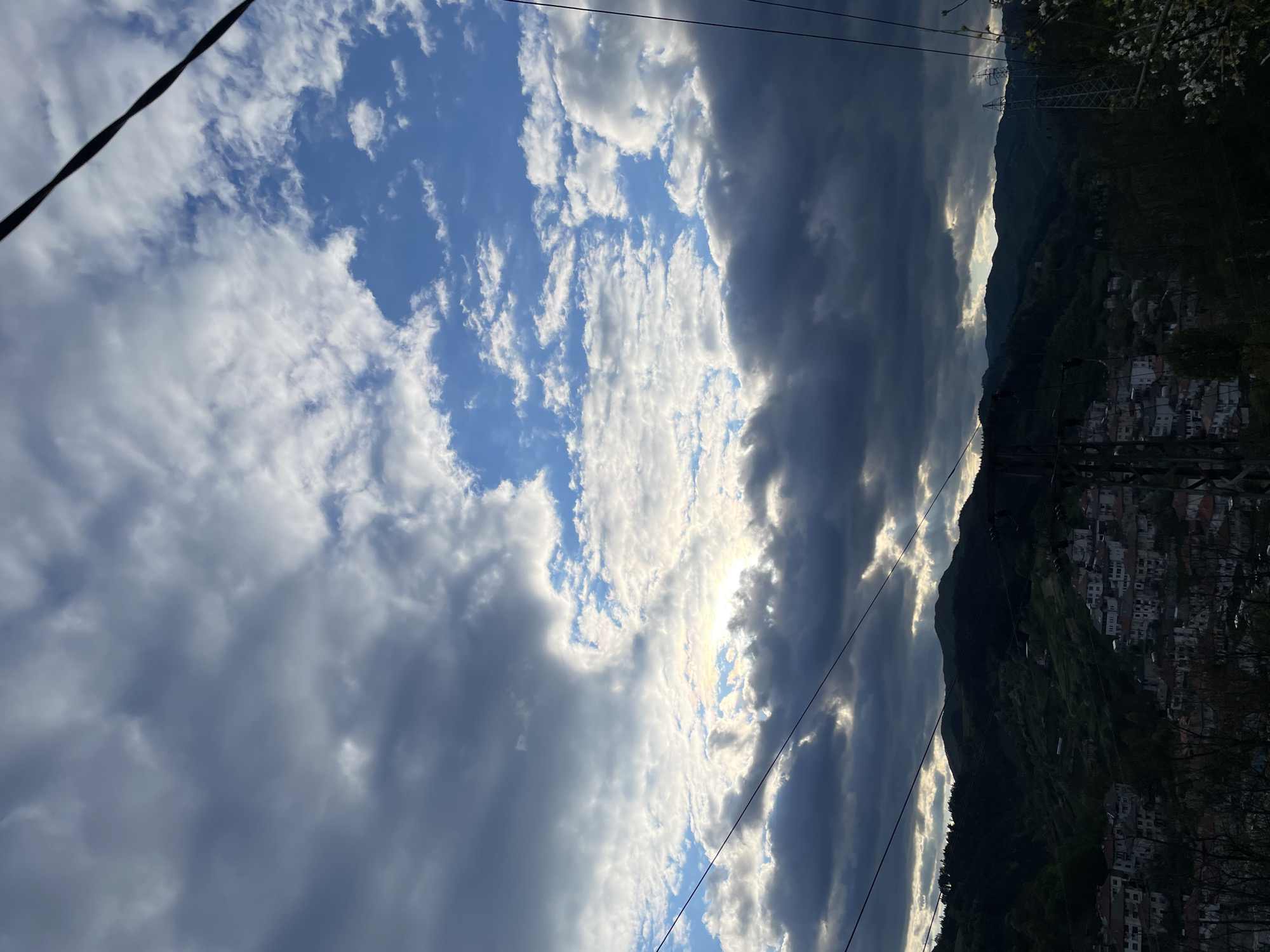 The image captures a view of Zlatograd, Bulgaria, under a partially cloudy sky. Dominating the right side of the frame is a mountain range covered in green vegetation and dotted with residential buildings. The town, with its white-walled structures, appears to be built on the slopes. In the sky, there's a mix of bright blue patches and dense, dark clouds, with sunlight filtering through the clouds. Power lines are visible, cutting across the sky.