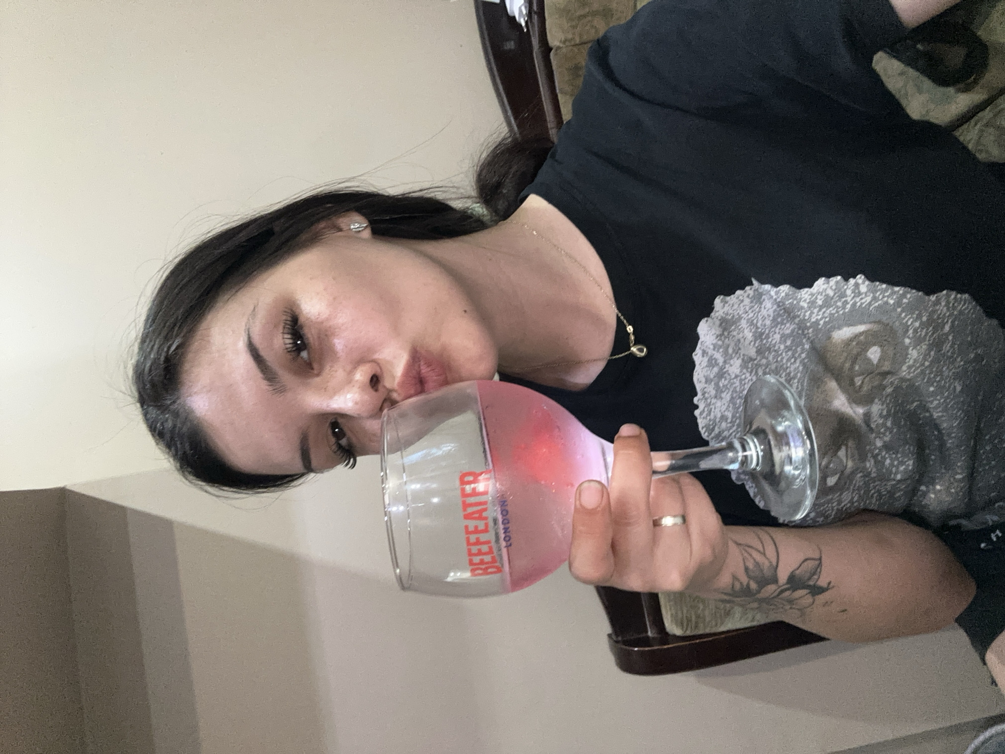 In a close-up, selfie-style shot, a young woman with dark hair pulled back and makeup, including false eyelashes and freckles, holds a large wine glass filled with a pink beverage. The glass, bearing the red "BEEFEATER LONDON" logo, is positioned in front of her face. She is puckering her lips as if to kiss the glass. She wears a black t-shirt with a grey, textured graphic on the front and a delicate gold necklace. A ring is visible on her left ring finger, and a tattoo of a flower with leaves is on her left forearm. The background is an indoor setting with a cream-colored wall and a dark wooden chair. The scene suggests a casual moment, possibly at home or a restaurant. No specific location in Sliven, Bulgaria can be determined from the image.