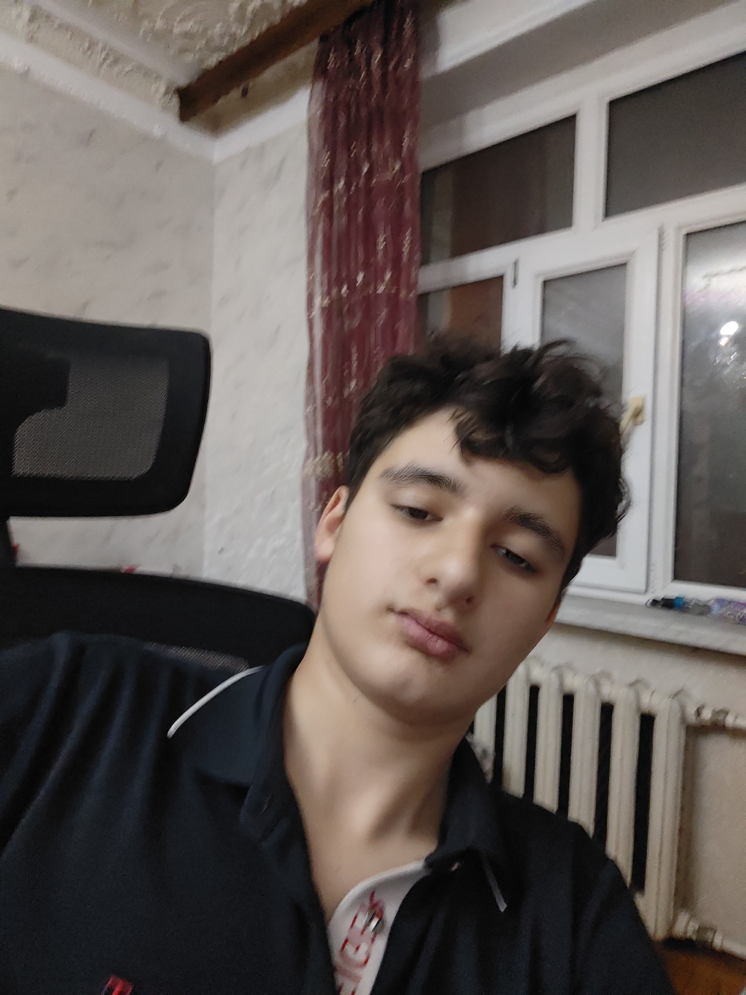 The image depicts a young male, appearing to be in his late teens or early twenties, seated in a black office chair. He is facing the camera and is partially visible from the shoulders up. He has dark, curly hair and is wearing a dark blue collared shirt with a white trim around the collar. The background shows a room interior with a textured wallpaper, a wooden beam near the ceiling, and a window with white frames and a burgundy curtain. A radiator is visible below the window. The scene appears to be an indoor domestic setting. There is no indication of the location being in the city of Baku City, Azerbaijan.