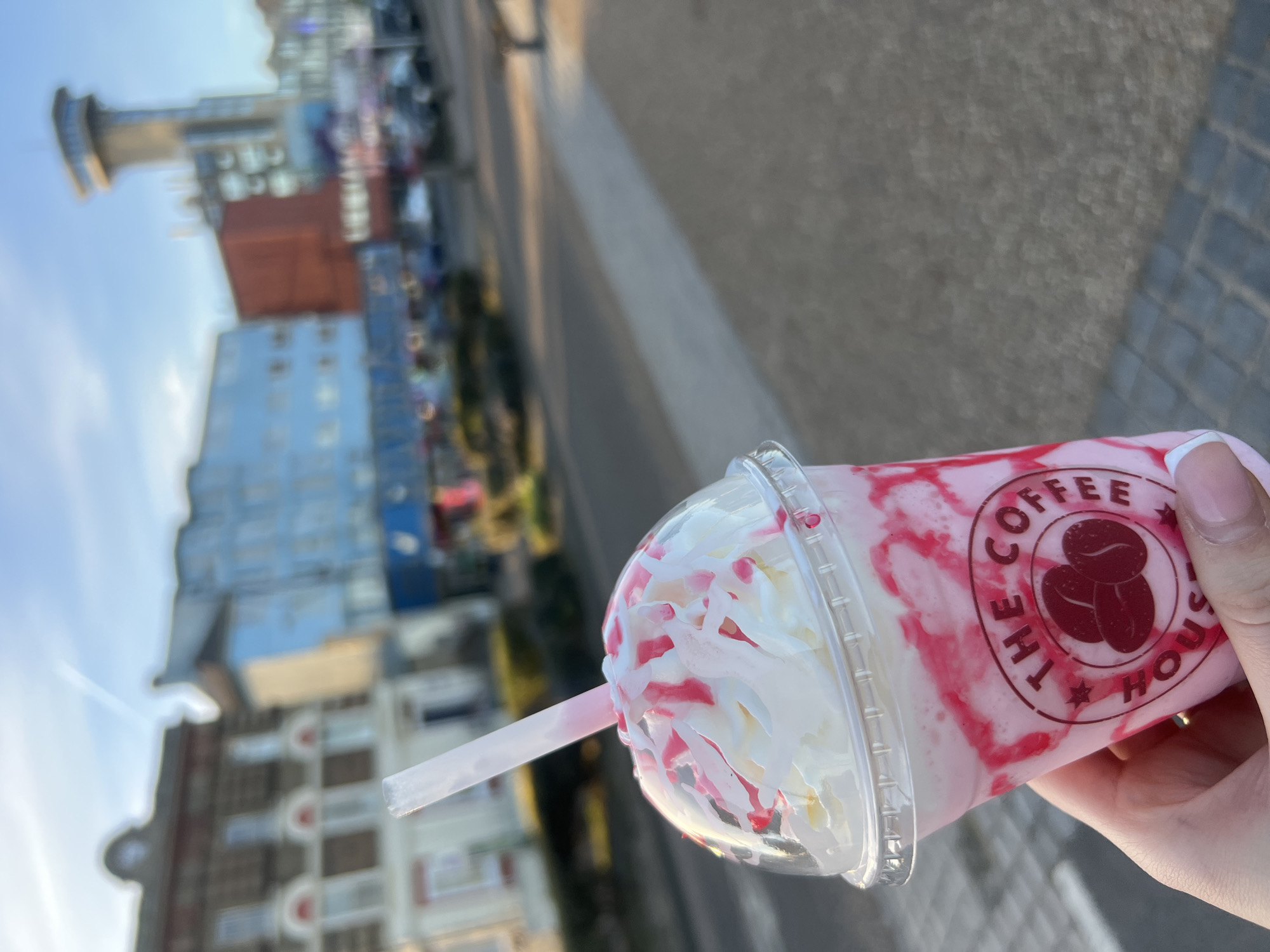 The image shows a person holding a pink and white milkshake or smoothie from "The Coffee House," a cafe chain. The drink has whipped cream and is drizzled with pink syrup. The backdrop features a blurred view of the seaside town of Gorleston-on-Sea, United Kingdom, with buildings and a tall tower visible against a partly cloudy sky. A sidewalk is also partially visible.
